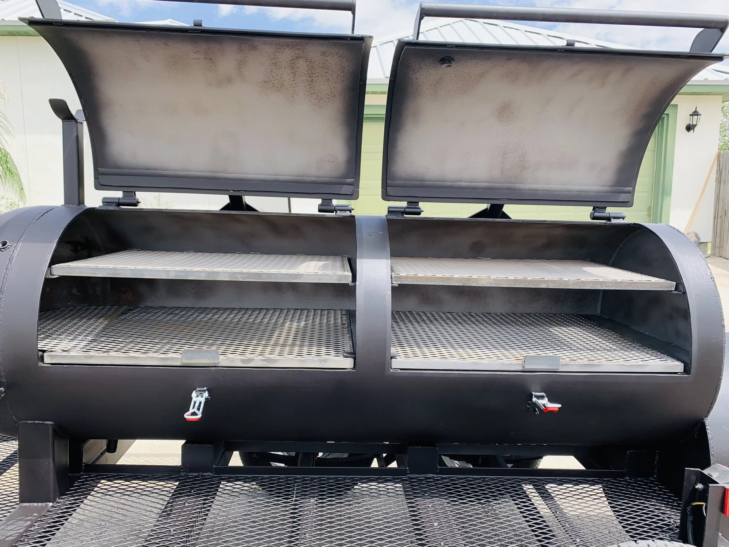 A close-up view of a black outdoor grill with two open lids, showing the cooking grates and interior. The grill is situated outdoors in front of a building with a green door and exterior light fixtures. The sky is partly cloudy.