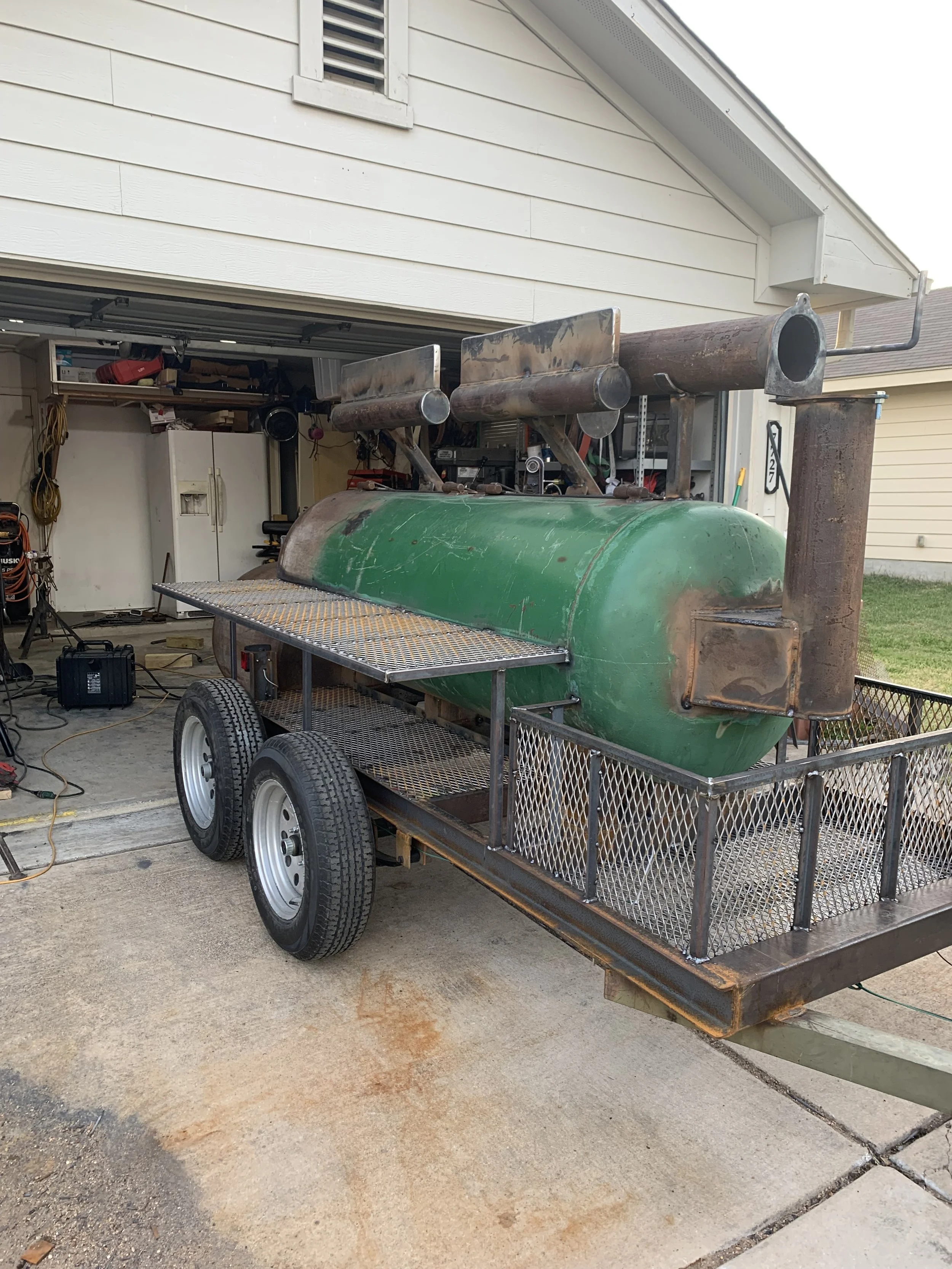 A flatbed trailer with a green propane tank attached, parked in a driveway near a garage with some tools and storage inside.