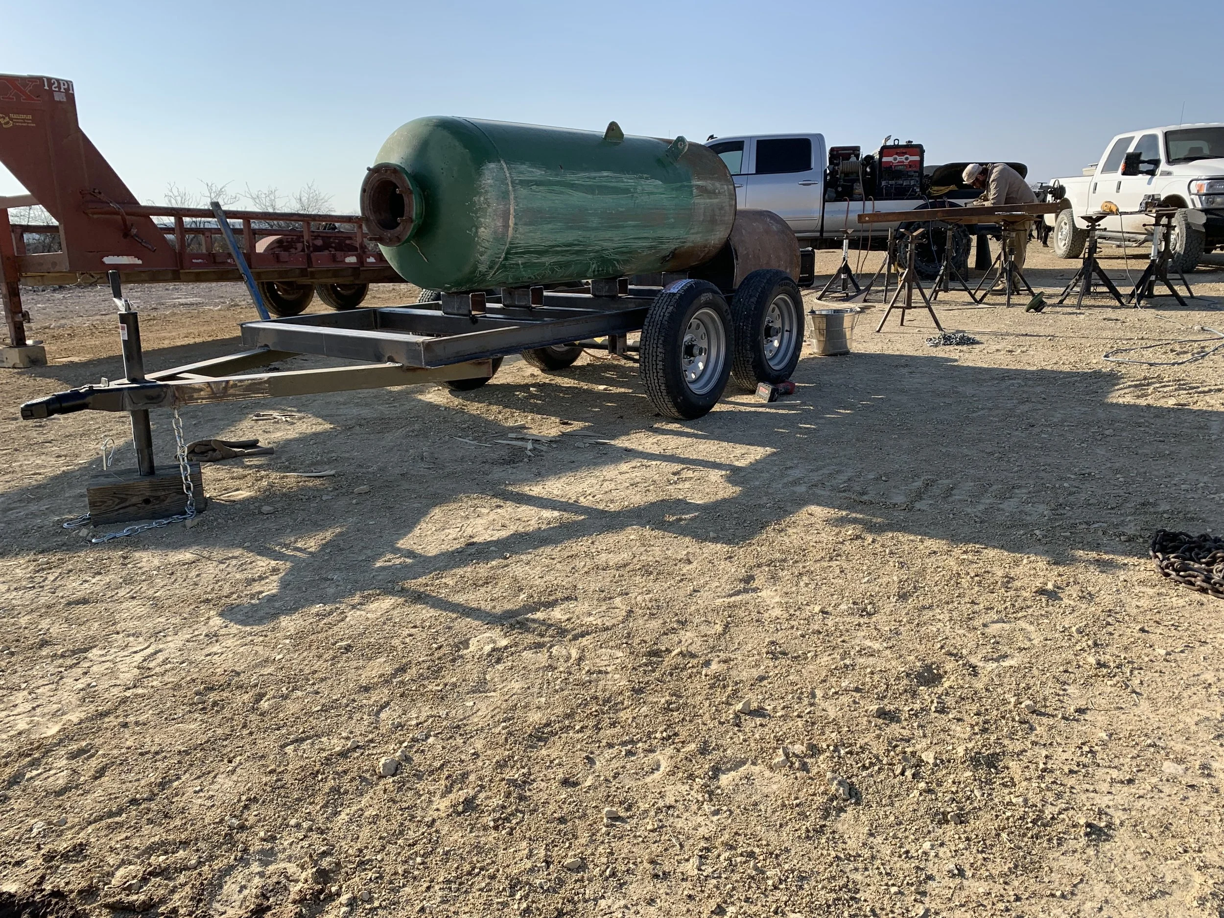 A large green cylindrical tank strapped onto a wheeled trailer on a dirt lot. Two trucks and a person working with equipment are in the background.