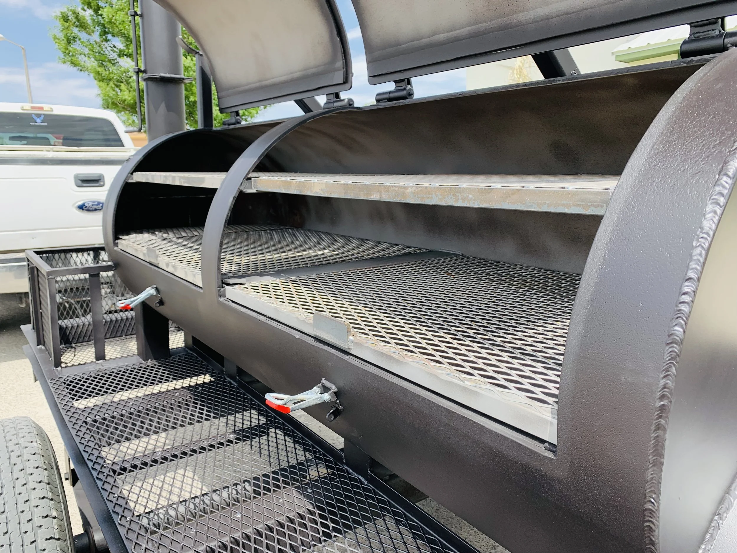 A large metal smoker with multiple shelves, situated outdoors in a parking lot, with a white Ford truck and a green tree in the background.