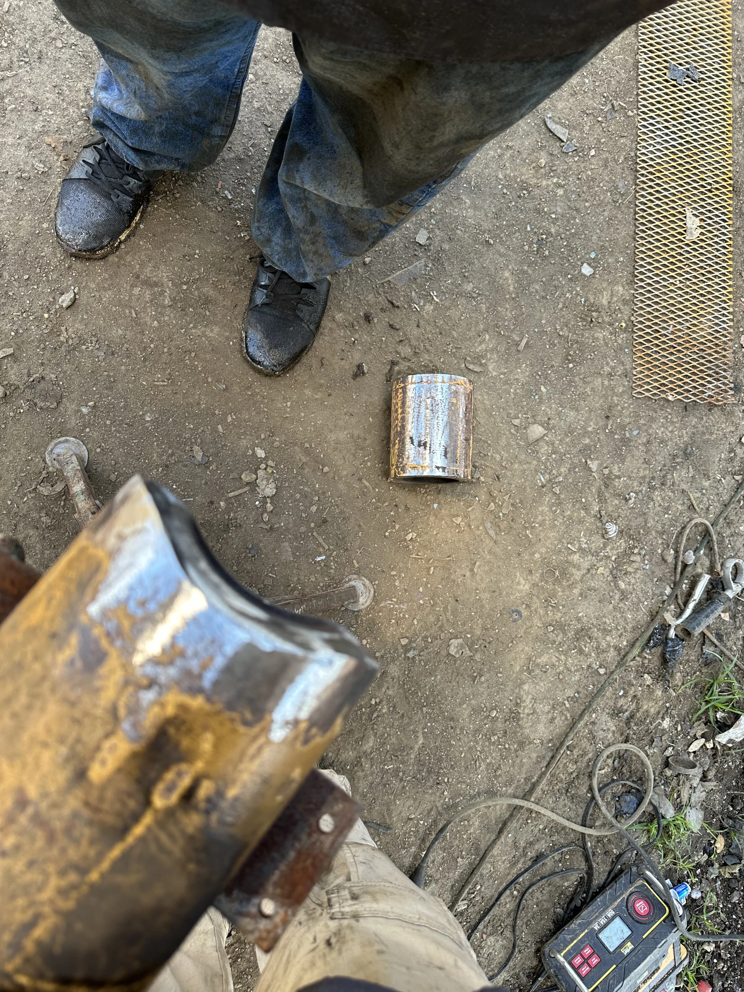 First-person view of a person in work clothes and sneakers standing on a dirt ground, with an electric device and a rusty metal pipe nearby, and tools on the ground.