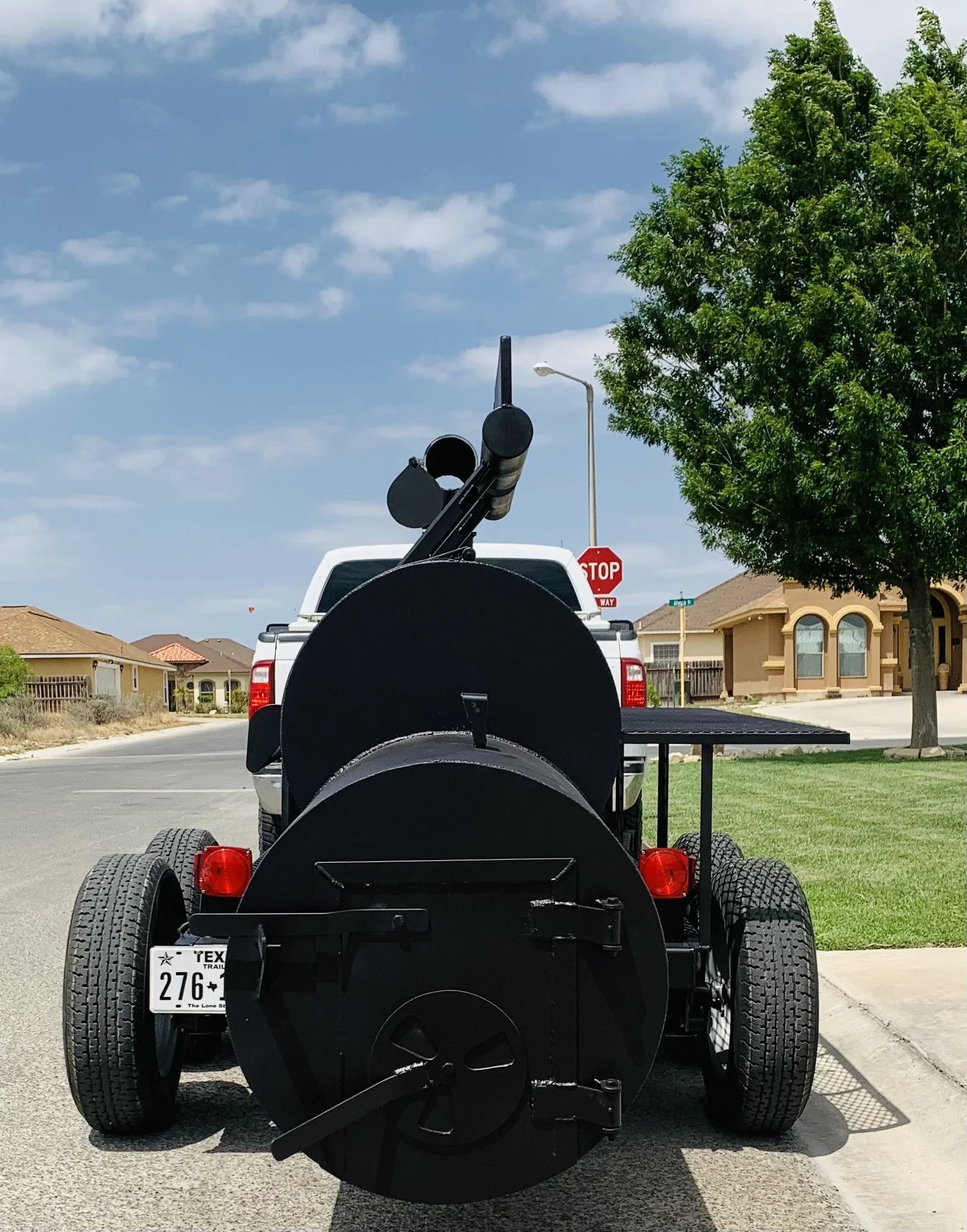 A truck with a large machine or equipment mounted on the back, parked on a residential street. The equipment appears to be a large, black, industrial or construction device with multiple components, including large black tires and a trailer hitch. Th