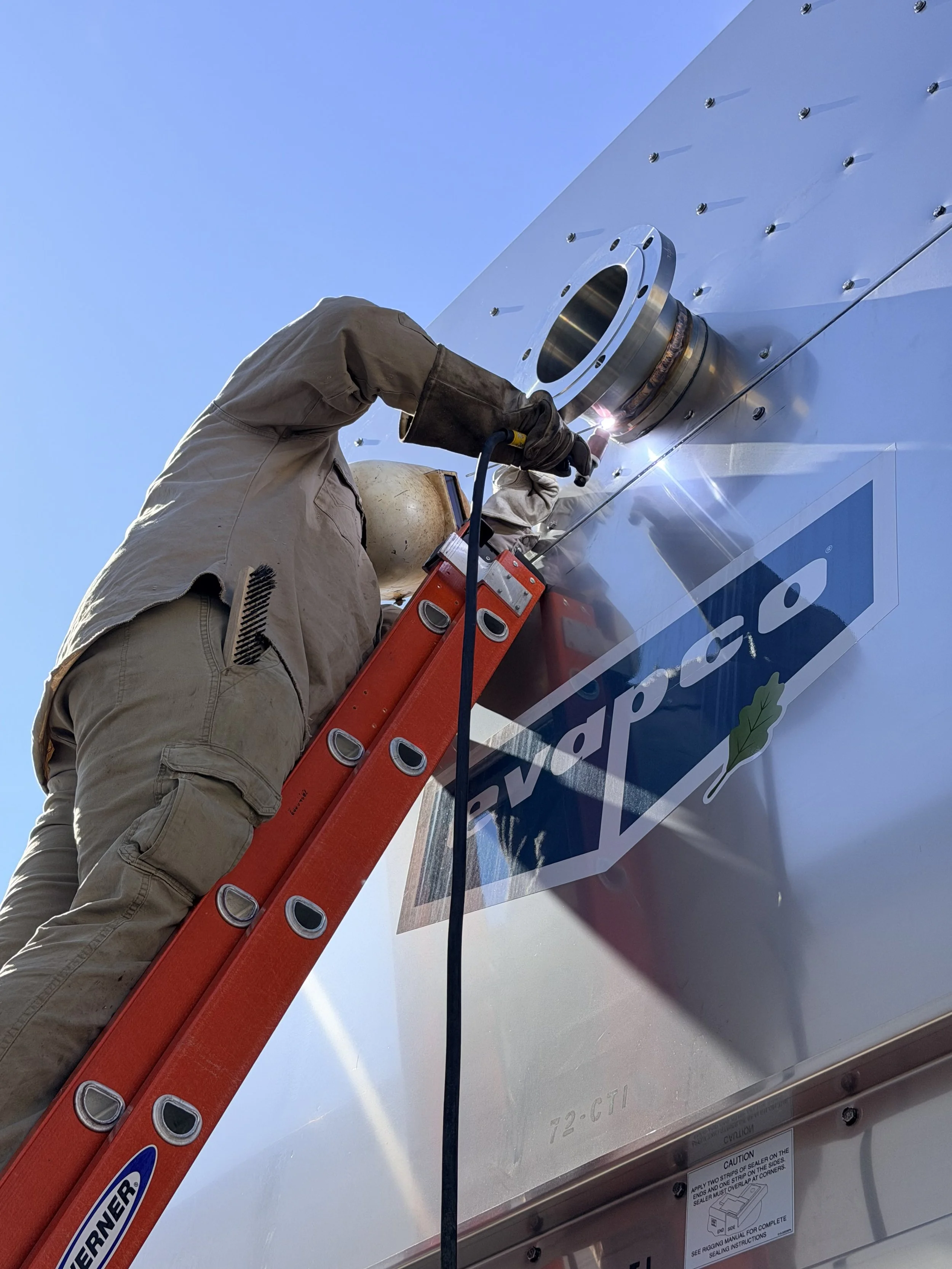 Technician welding a metal component onto a large industrial machine or vehicle with a red ladder positioned against it.