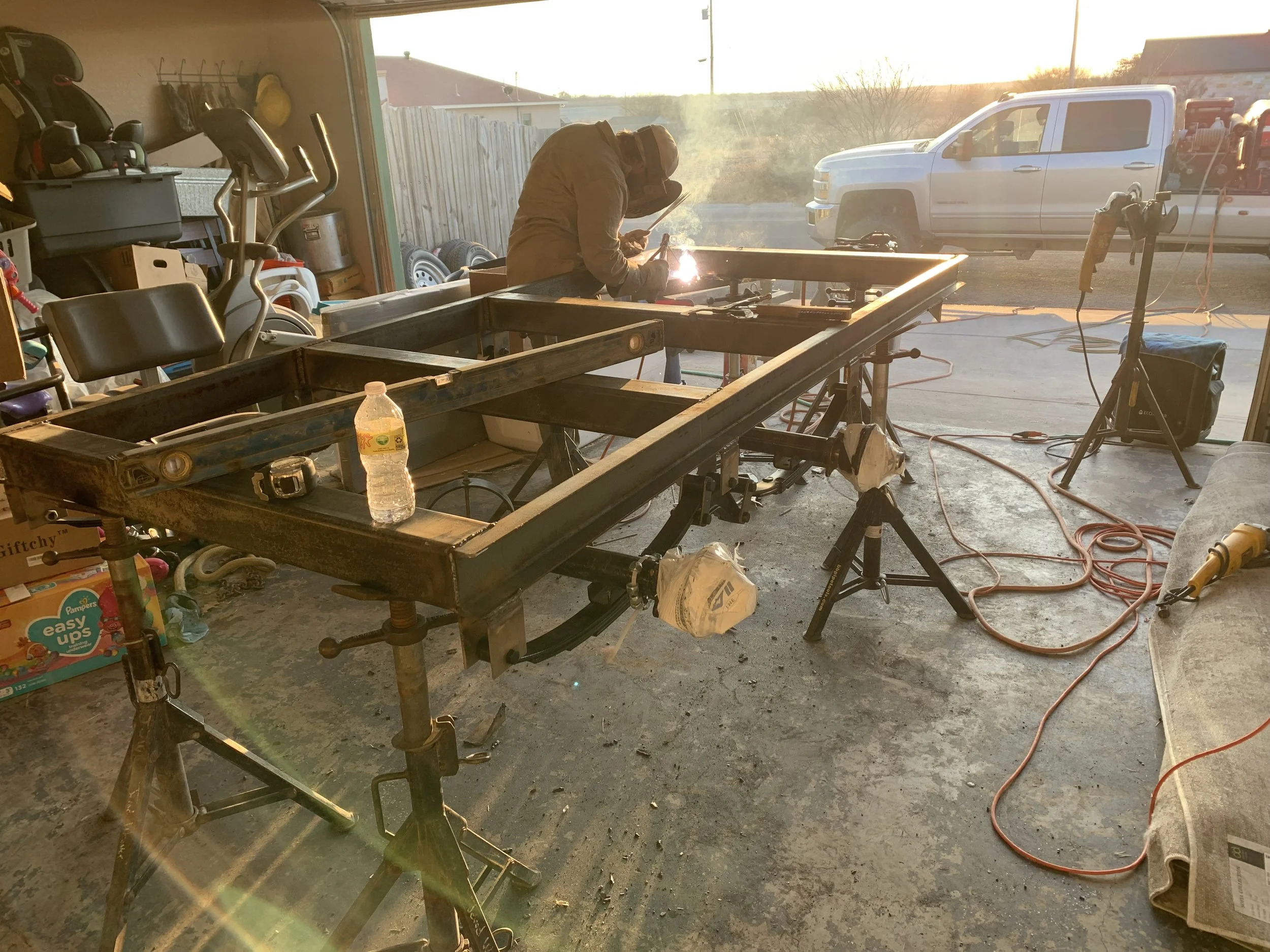 A person welding in a garage or workshop, with sunlight shining through an open door. There are various tools and equipment around, including a work table, a water bottle, and a stationary bike.