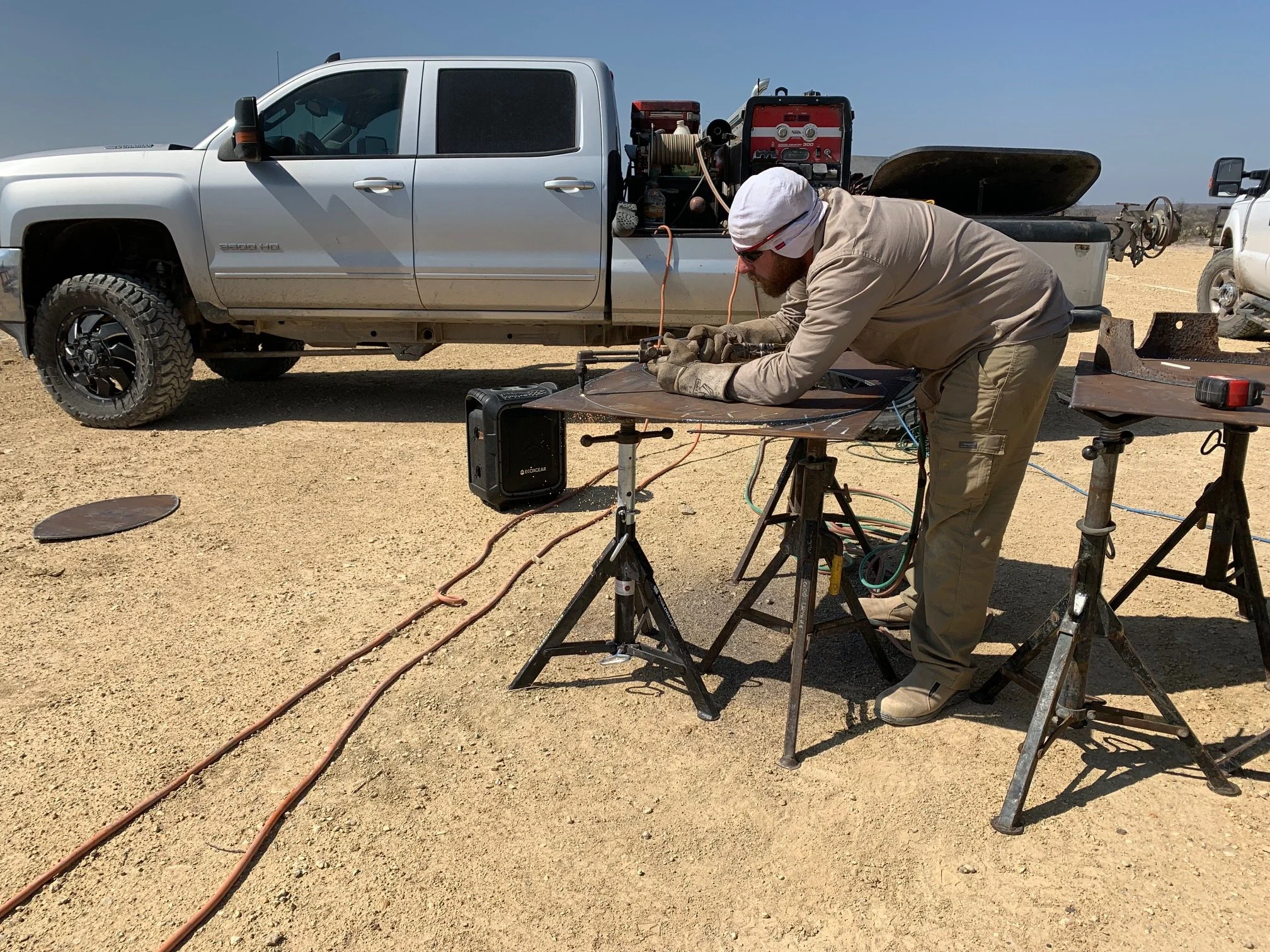 A man working on the underside of a pickup truck in a desert setting, with tools and equipment nearby.