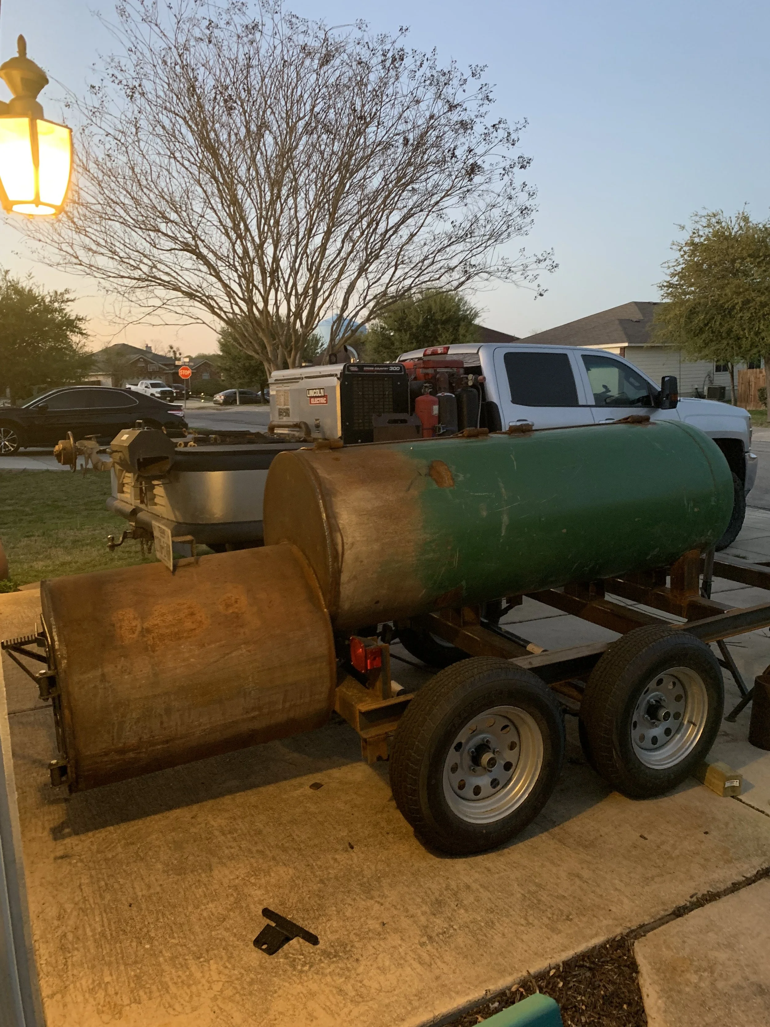 The image shows a suburban scene with a large, leafless tree in the background, a white pickup truck in the middle, and several pieces of equipment including a large green metal tank and a rusty brown barrel on a trailer in the foreground. There is a