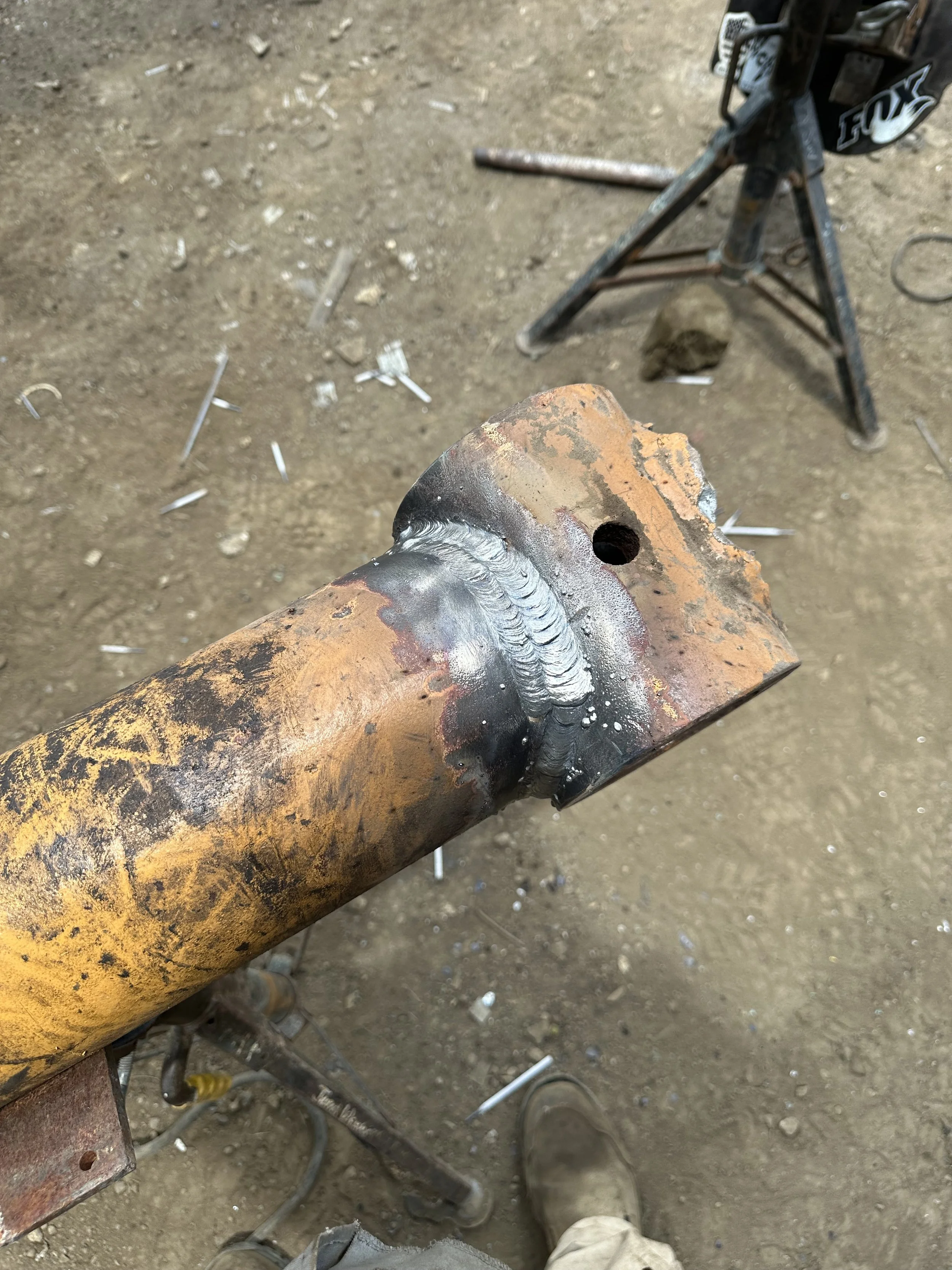 Close-up of a rusty metal pipe with fresh weld bead along the joint with a round hole, on a dirt ground with scattered metal shavings and tools nearby.