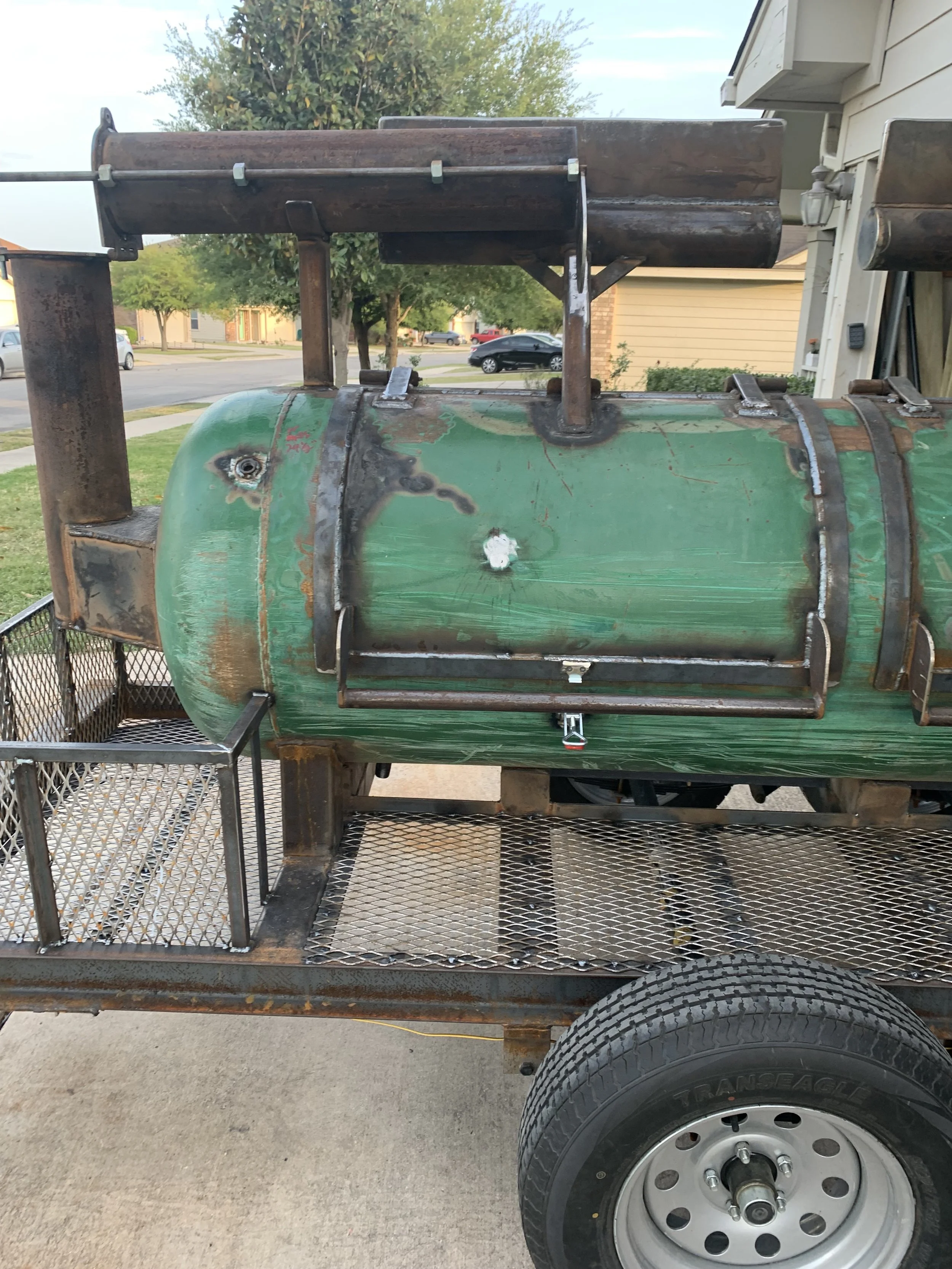 Front view of a weathered green utility trailer with a black metal frame, metal mesh sides, and a single tire, parked outdoors on a concrete driveway with residential houses and trees in the background.