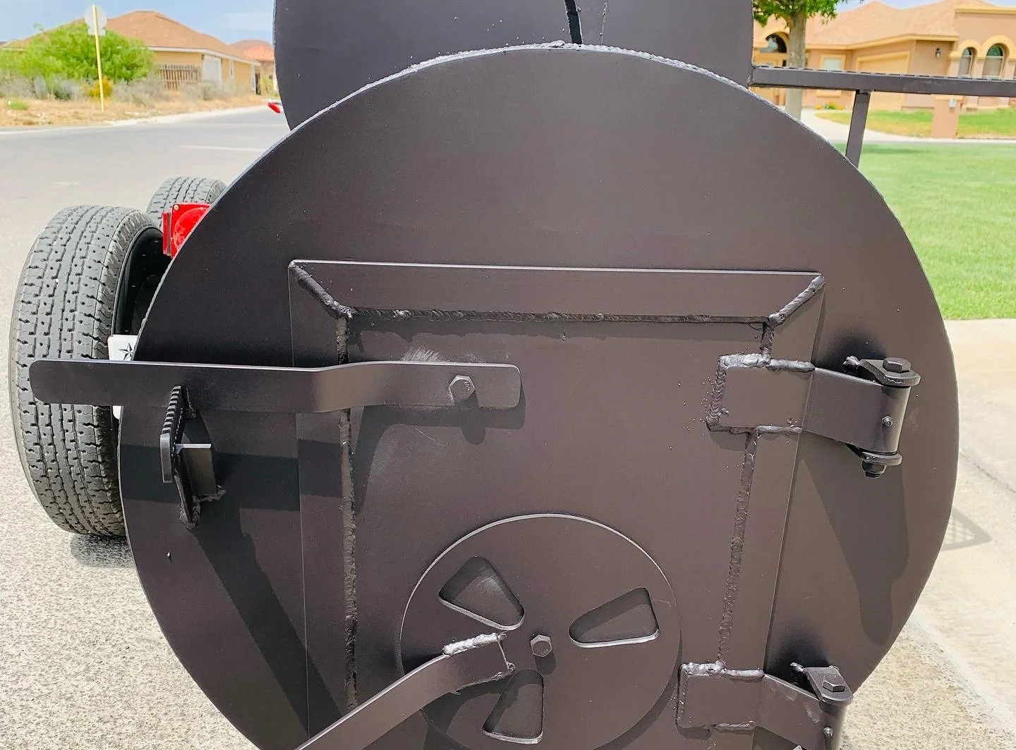 Close-up of a large, black, circular industrial machine or equipment with welded metal parts, located outdoors on a concrete surface, with a residential neighborhood and grass in the background.
