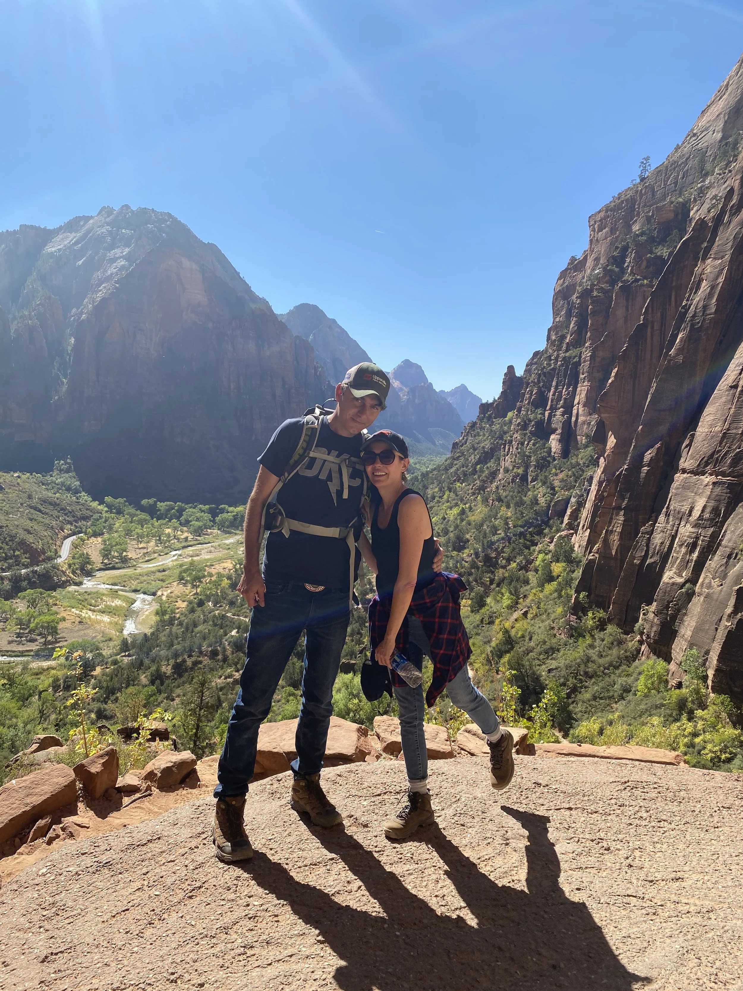 A man and woman smiling and posing on a rocky trail in a canyon with towering red and brown cliffs and a bright blue sky with sunlight.