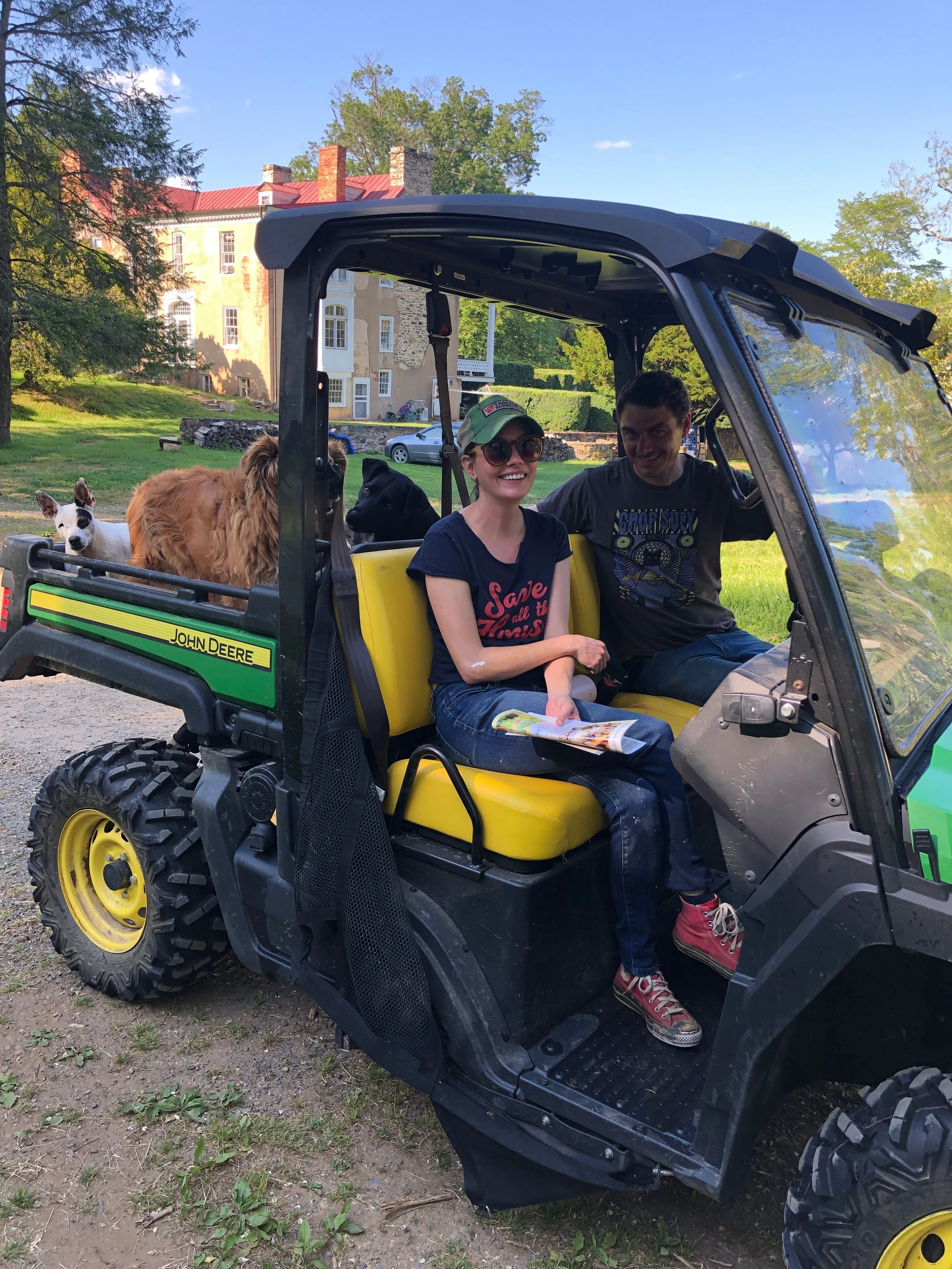 A man and a woman sitting in a yellow and black John Deere utility vehicle with two dogs, one large and brown and the other small and white, on the back. They are outdoors on a grassy area with trees and a large building in the background.