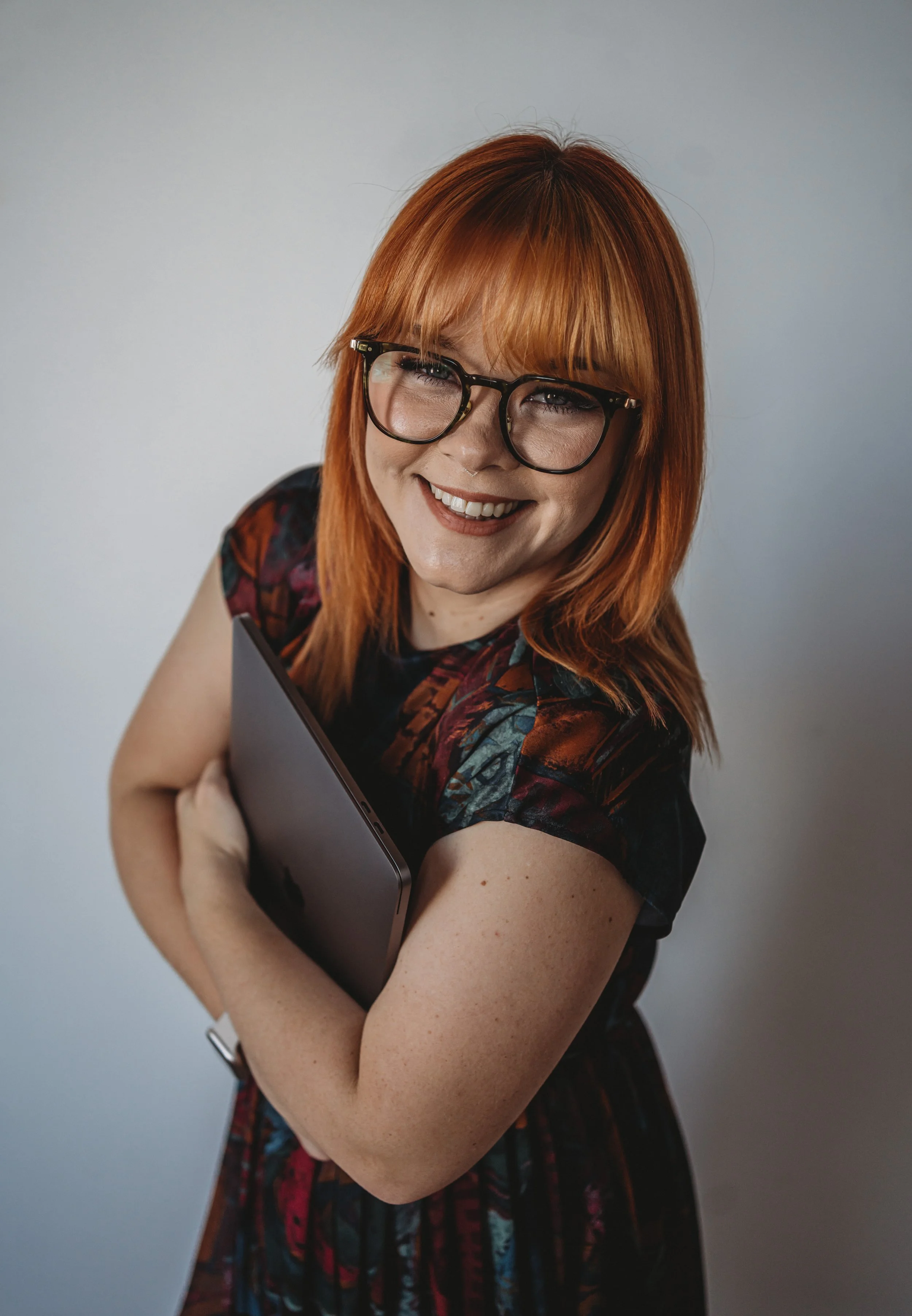 A woman with red hair and glasses smiling, holding a closed laptop or tablet, standing against a gray background.