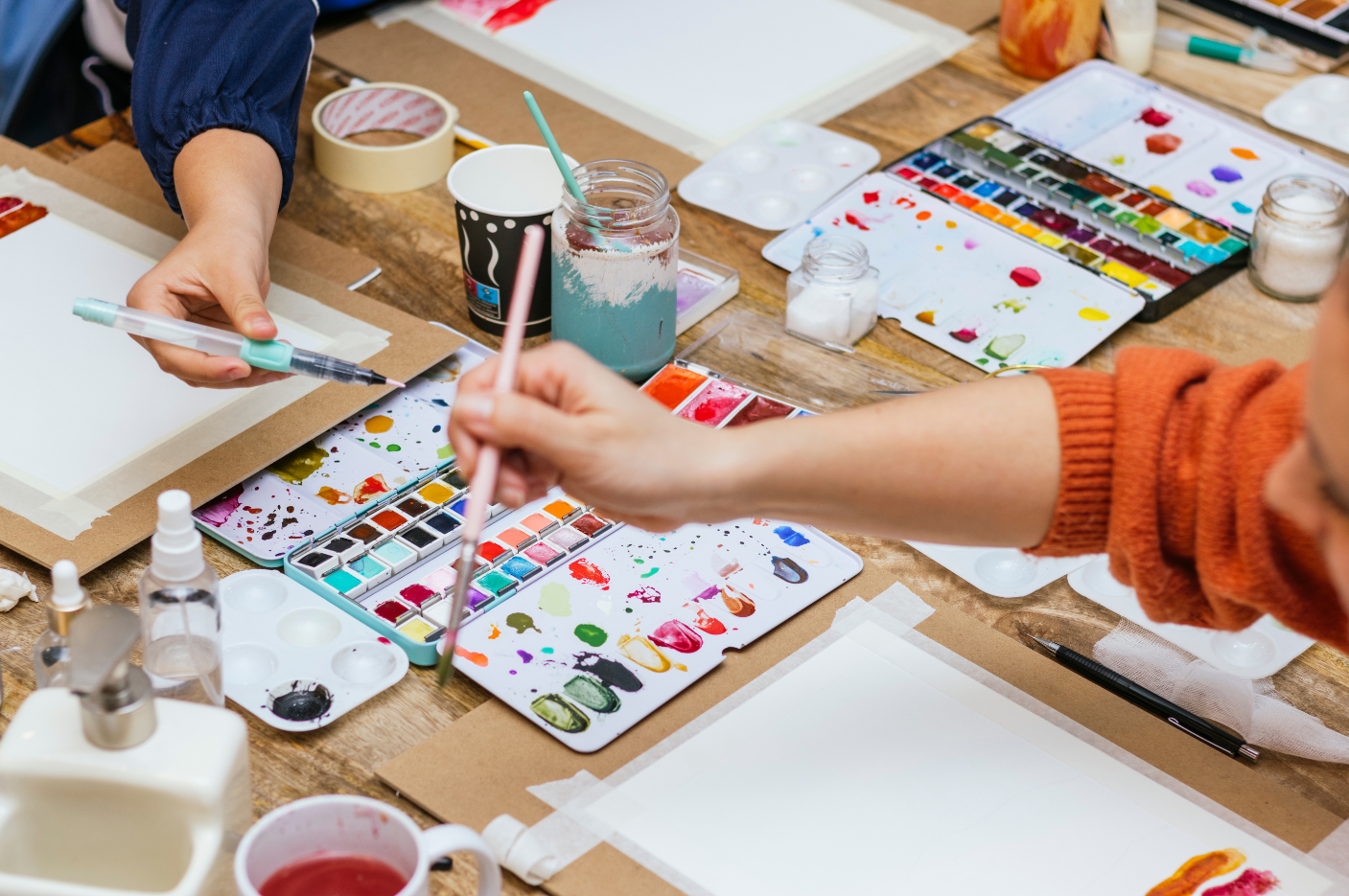 Artists painting with watercolor brushes at a workshop table surrounded by palettes, paint supplies, and taped watercolor paper