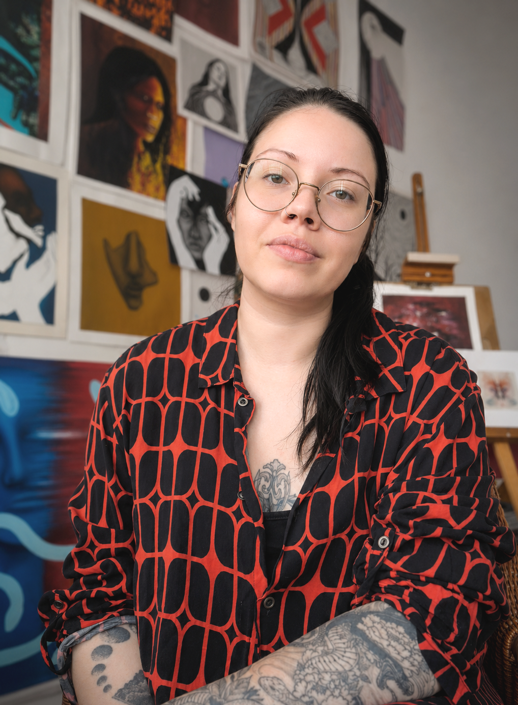 Tattooed woman wearing round glasses and a red-and-black patterned shirt in an art studio surrounded by framed artwork
