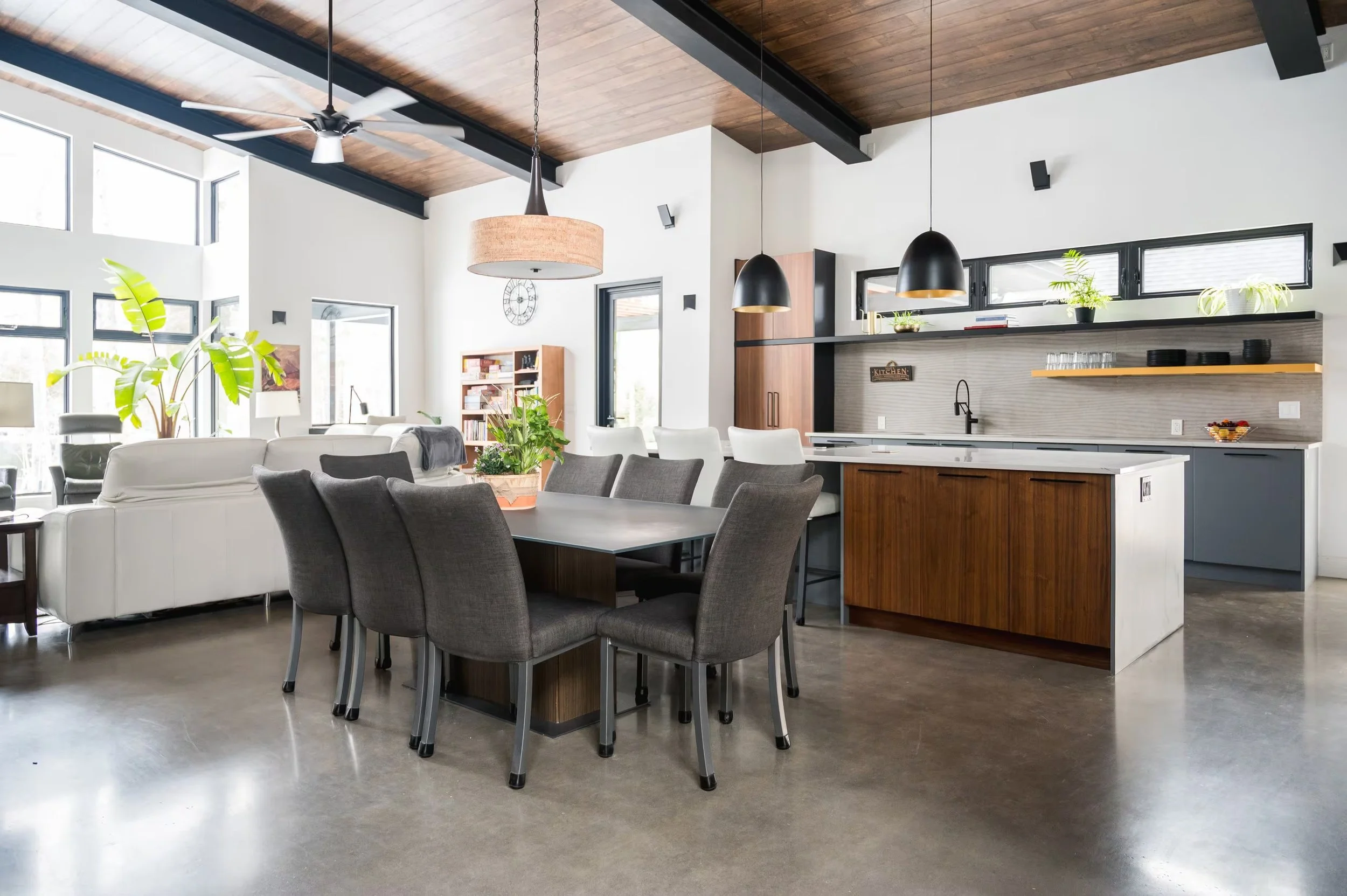 Open-concept living room and kitchen with high ceiling, wooden beams, large windows, modern furniture, plants, and pendant lights.