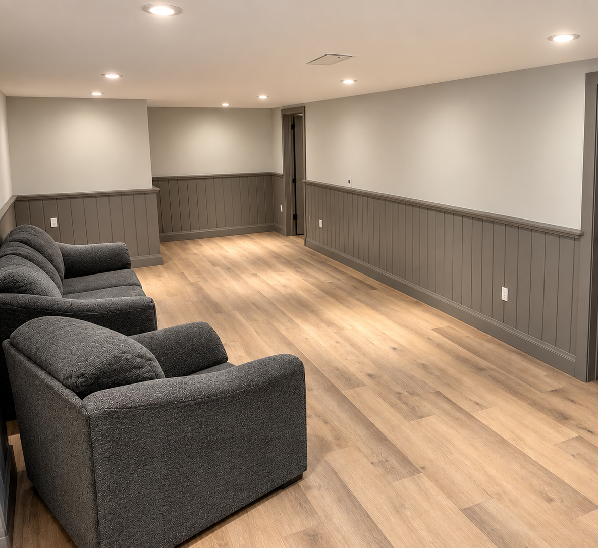 Empty basement room with wood flooring, gray walls with wood paneling, recessed lighting, and a row of gray upholstered armchairs.