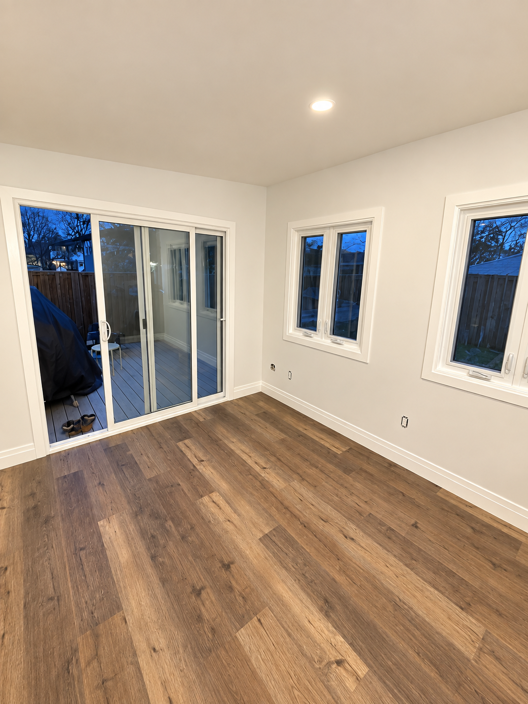 Empty room with wood flooring, white walls, three windows, and a sliding glass door leading to a deck.