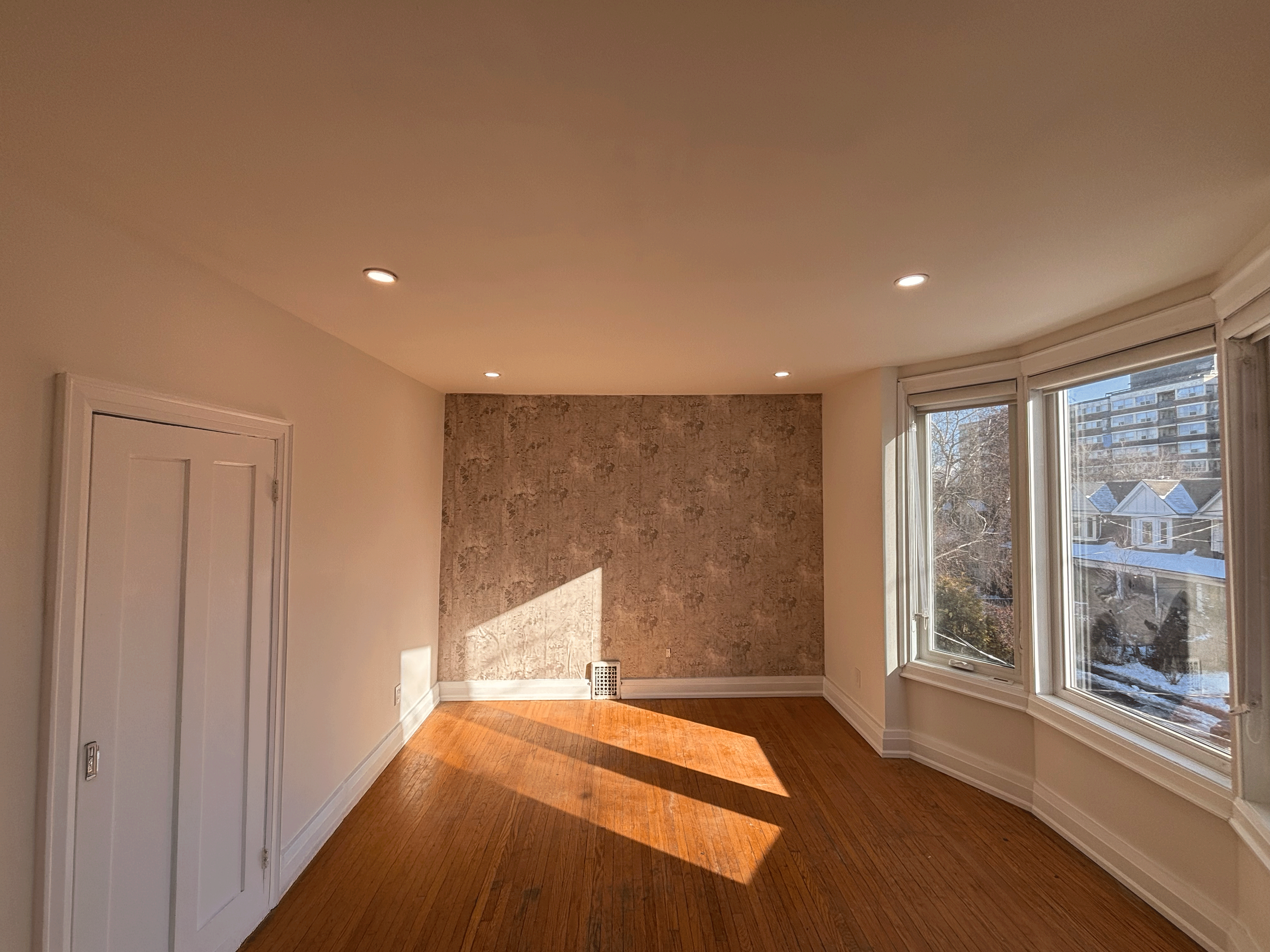 Empty room with hardwood floors, large windows letting in sunlight, a textured accent wall, and recessed lighting.