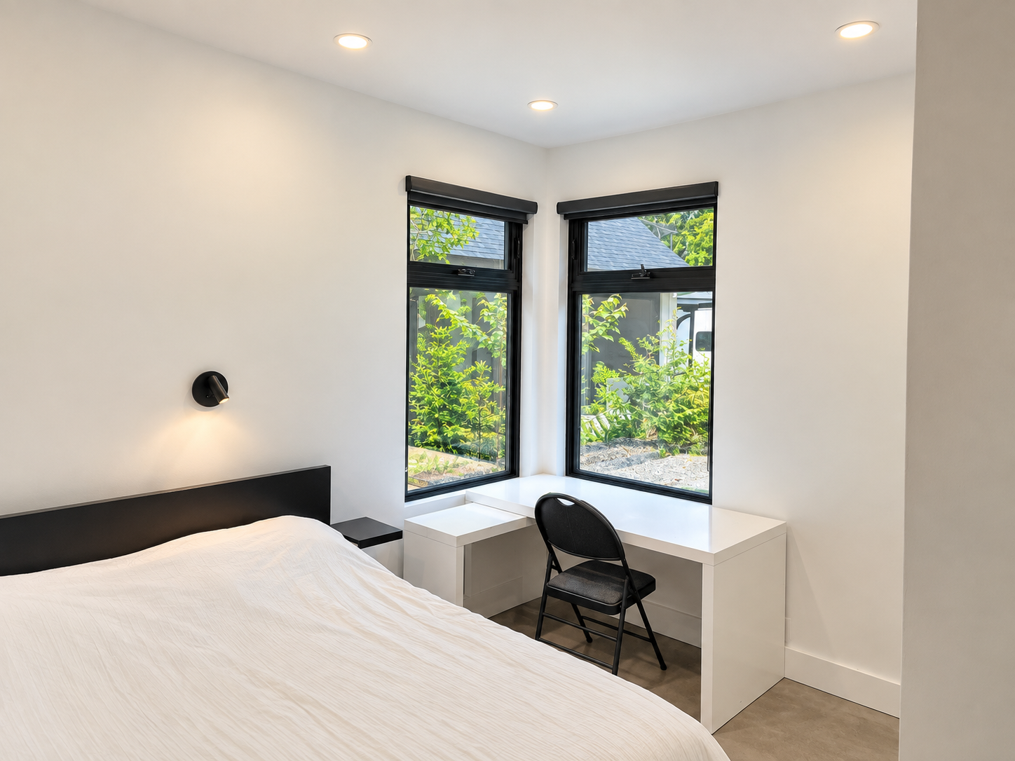 A minimalist bedroom with a bed, black headboard, two black-framed windows with a view of green foliage, a white desk, and a black chair.