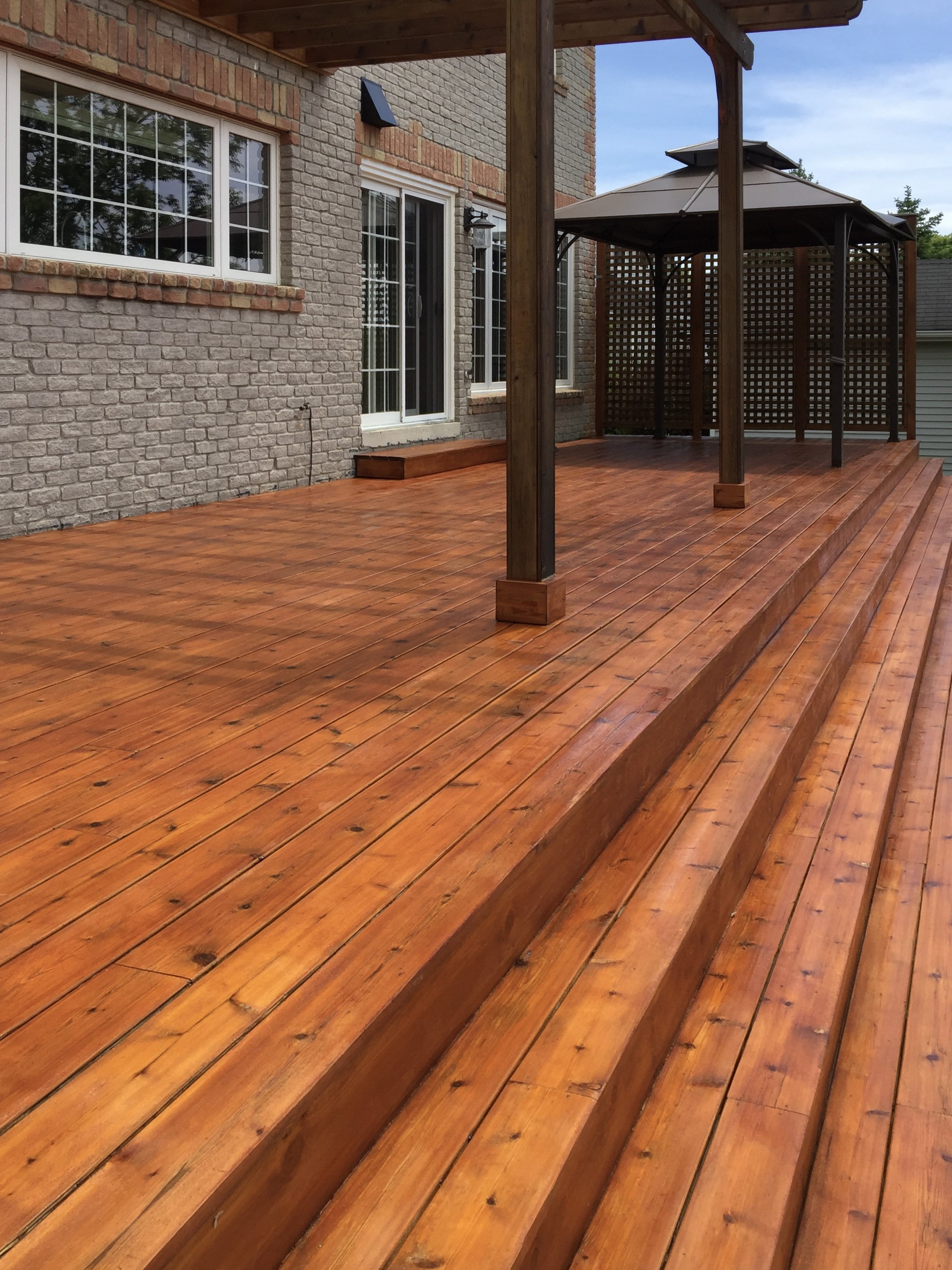 A wooden deck with stairs leading up to the house, a gazebo on the deck, and a brick house with white-framed windows and a sliding door.