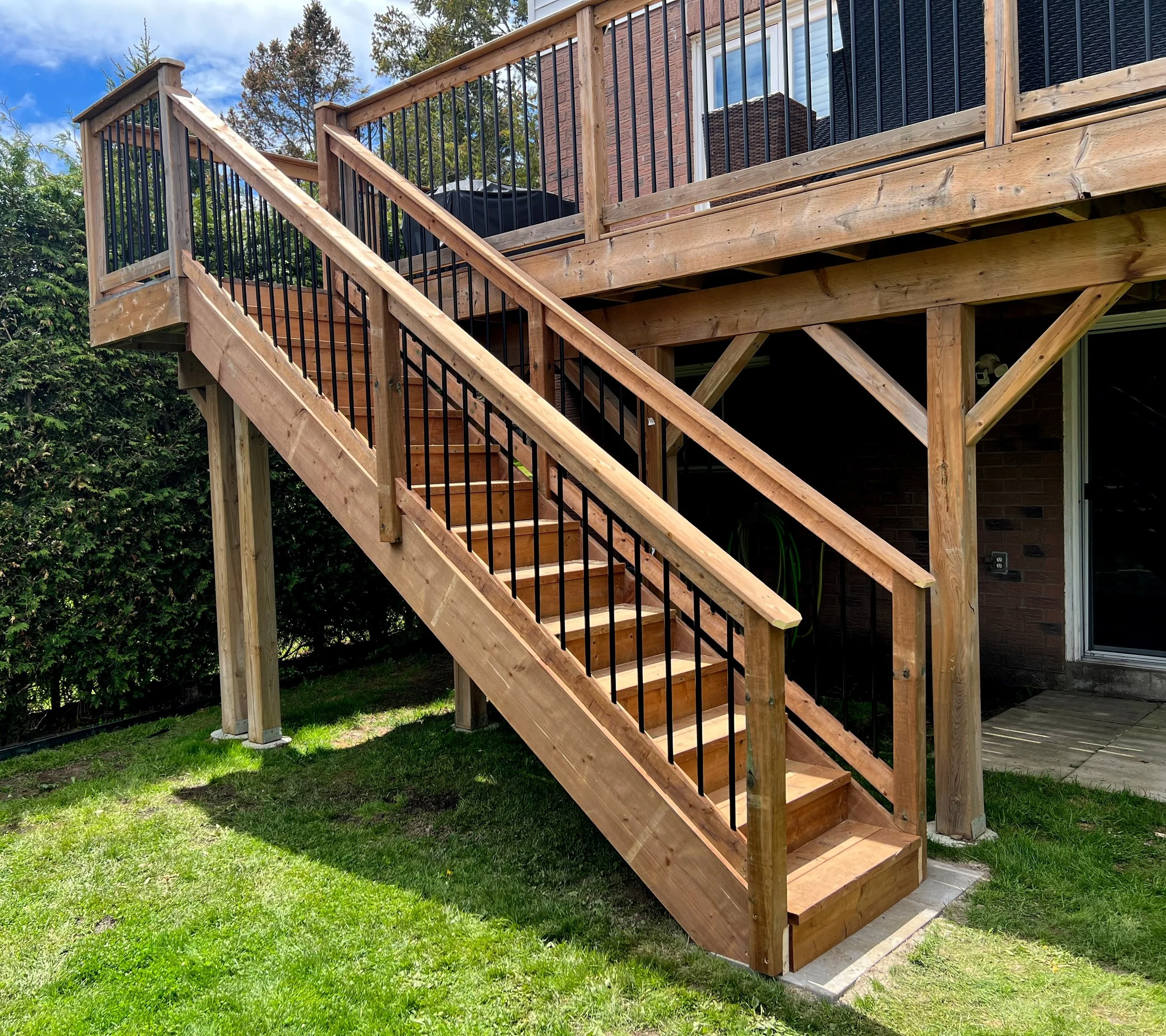 Wooden deck with black railing and stairs, attached to a brick house with sliding glass door, surrounded by trees and grass, with a cloudy sky.