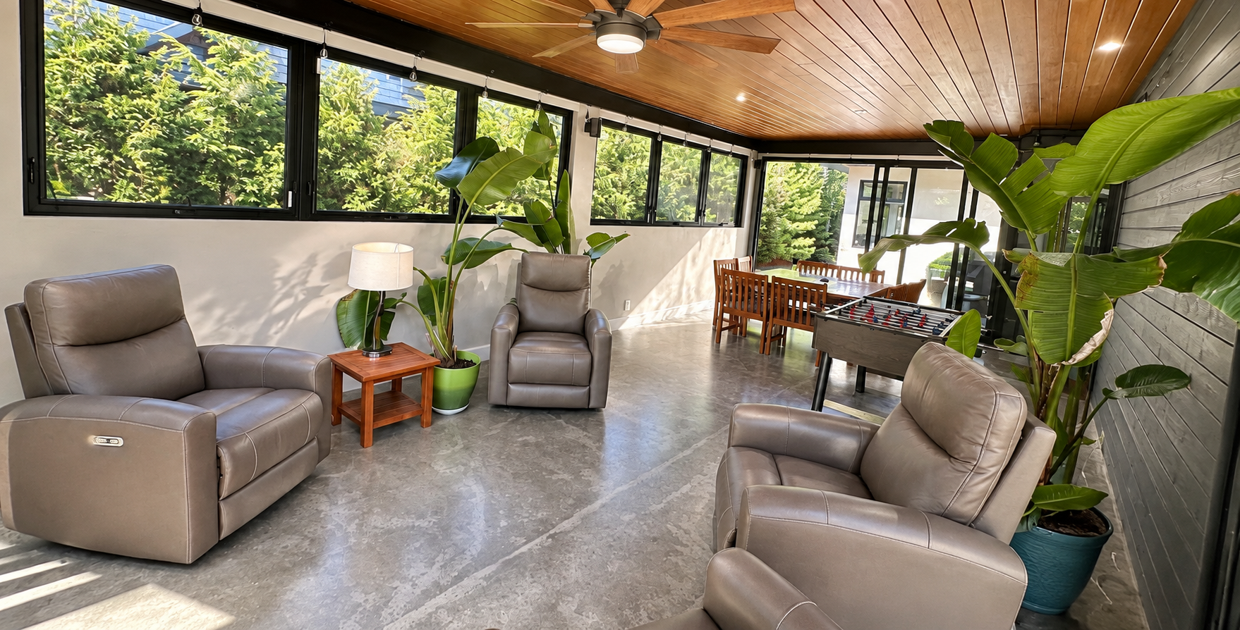 Indoor living space with leather armchairs, a wooden side table with a lamp, large green potted plants, a foosball table, and a dining table. Large windows let in natural light, revealing green trees outside. The ceiling is wooden with a ceiling fan.