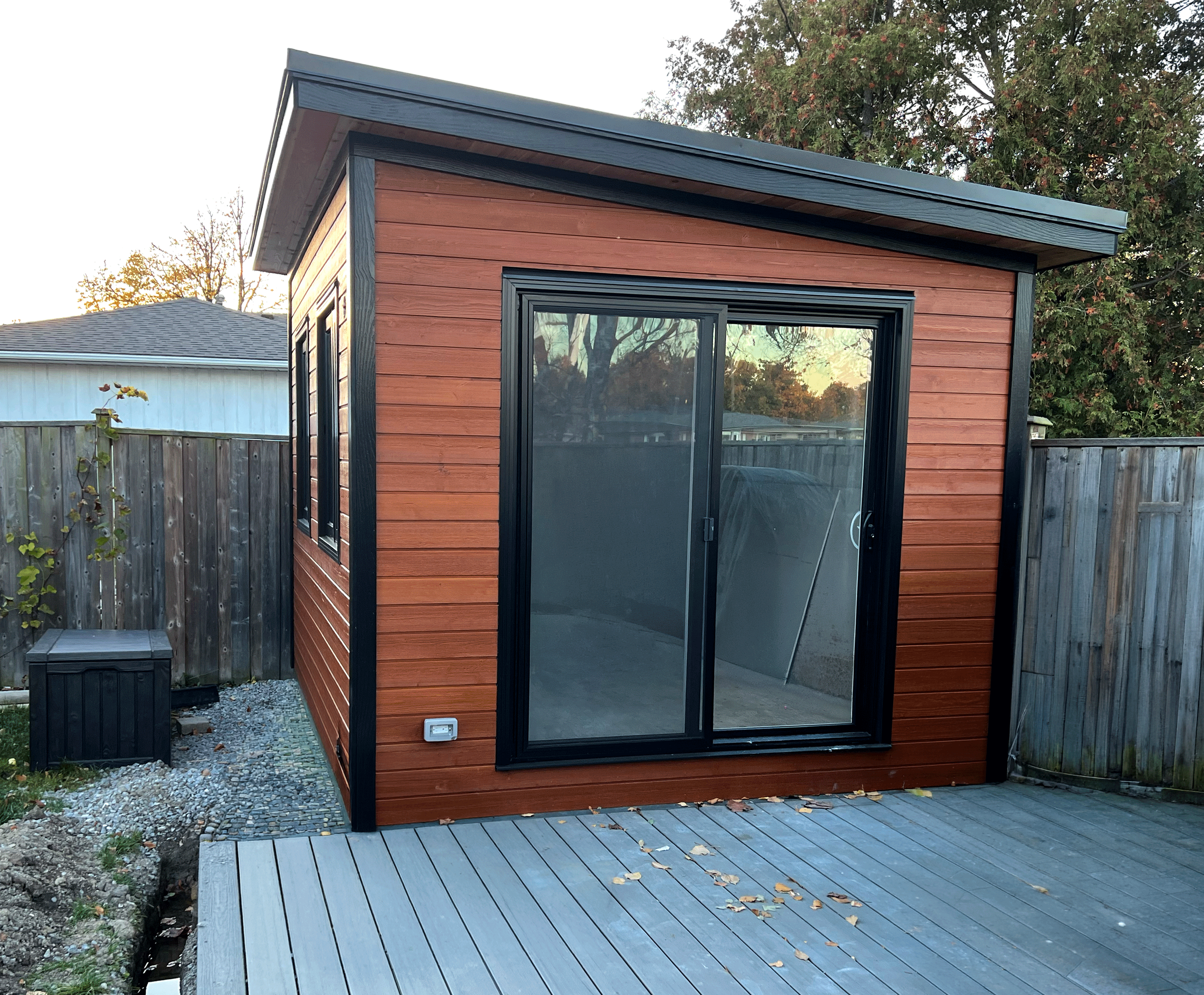 A small wooden building with a modern design, featuring horizontal reddish-brown wood siding, black framing around a large glass door, and a sloped roof, situated in a backyard with a wooden fence and a gravel path.