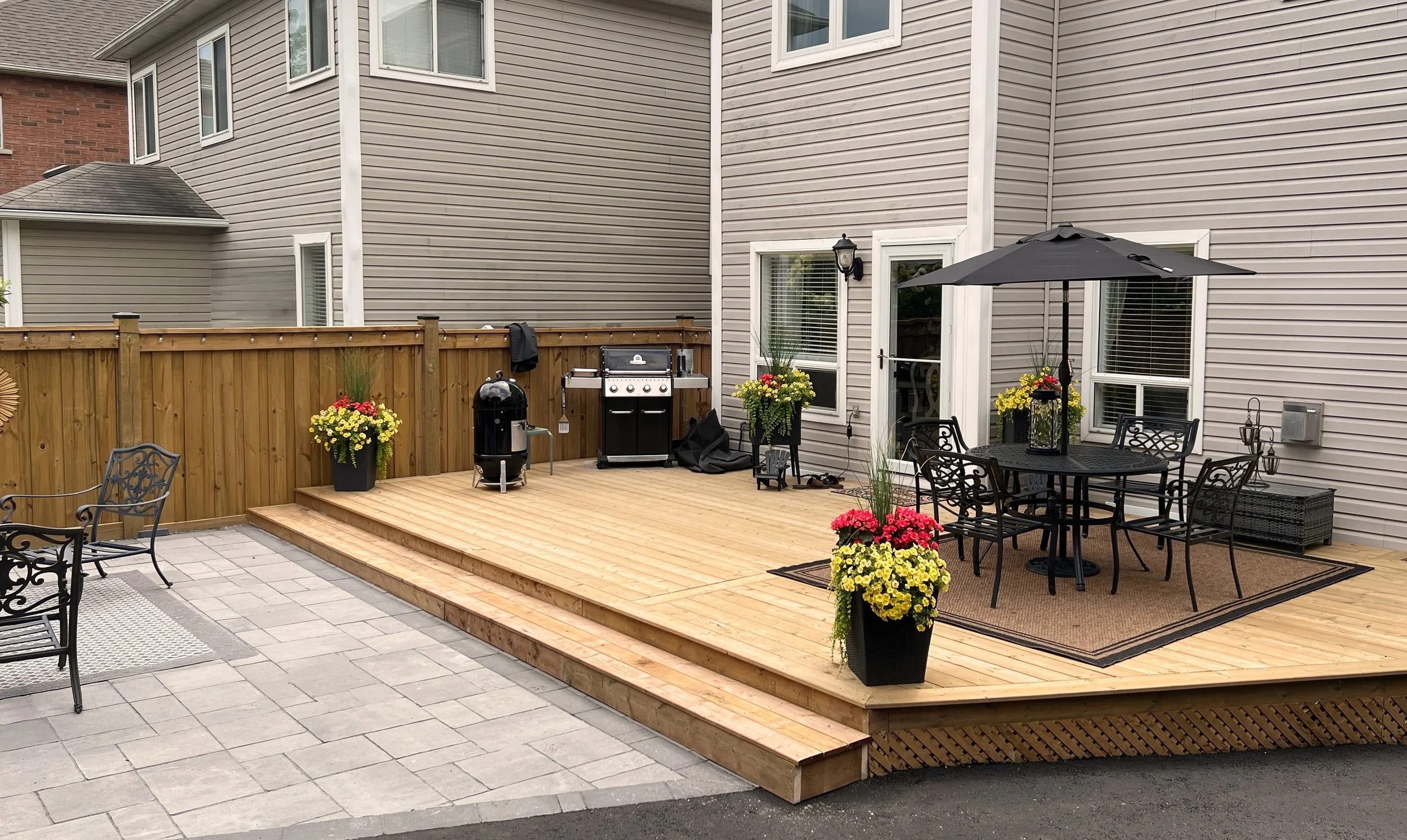 Backyard patio area with a wooden deck, black round table with chairs, large black umbrella, potted yellow and pink flowers, and a grill.