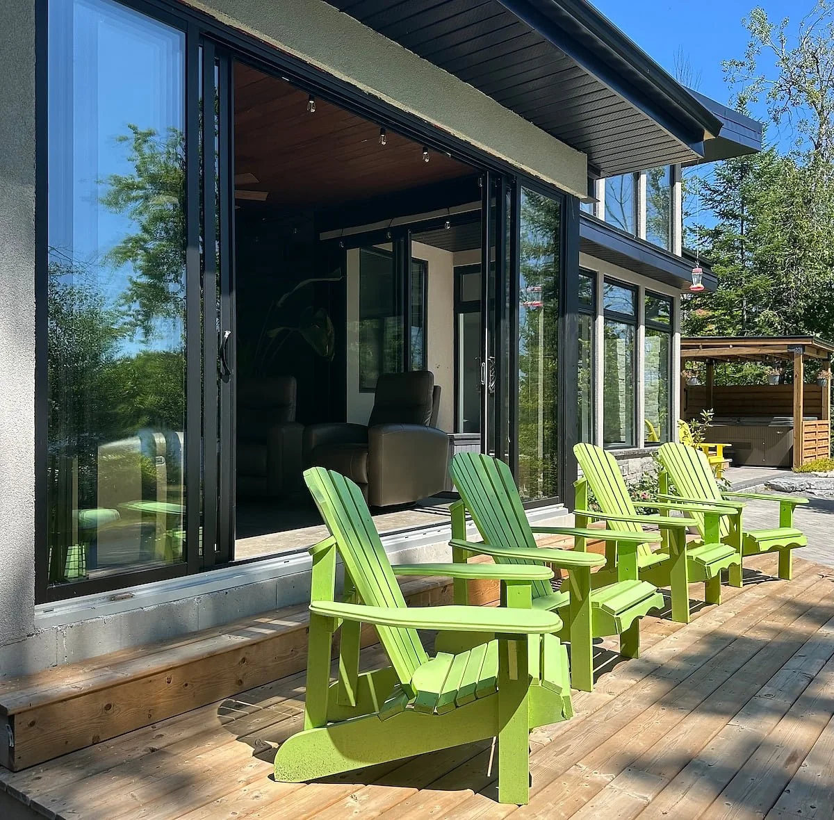 Outdoor wooden deck with four bright green Adirondack chairs. Behind, a modern house with large glass sliding doors shows a living room with black recliner chairs inside. To the right, a wooden gazebo with hot tub is visible, surrounded by trees and clear blue sky.