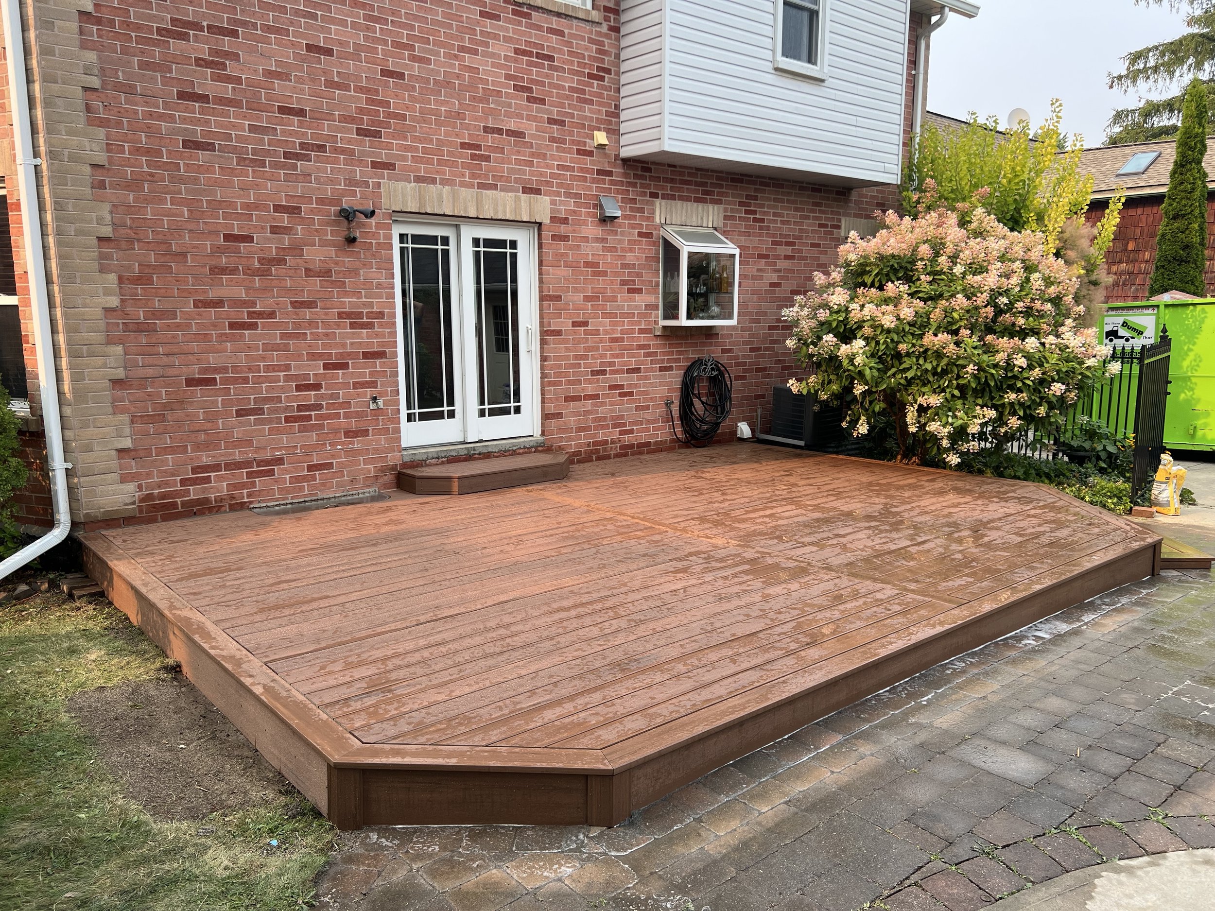 A backyard patio with a wooden deck, brick house exterior, white glass double door, a unique large flowering shrub, garden hose, and nearby green trash bins.