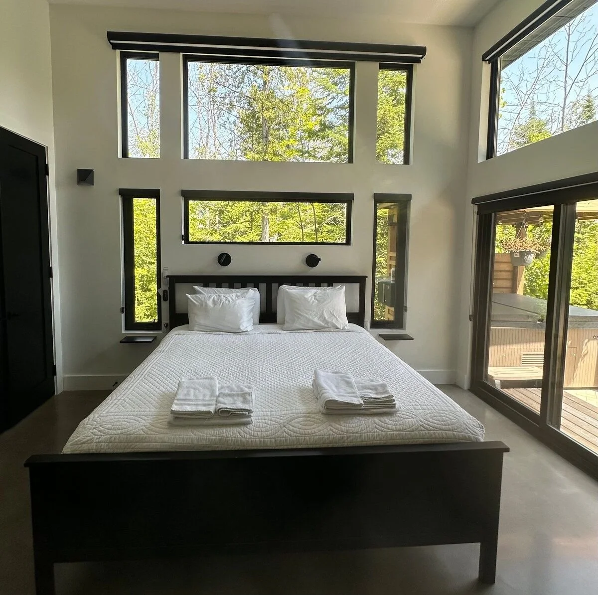 Modern bedroom with large windows showing trees outside, a black bed frame with white bedding, and towels on the bed, overlooking a deck.