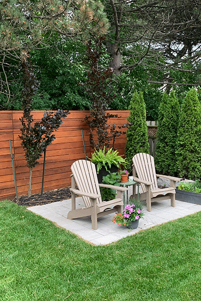 Two wooden Adirondack chairs with a small table between them on a paved patio in a backyard, surrounded by green grass, tall trees, and bushes, with potted flowers on the table and on the ground.