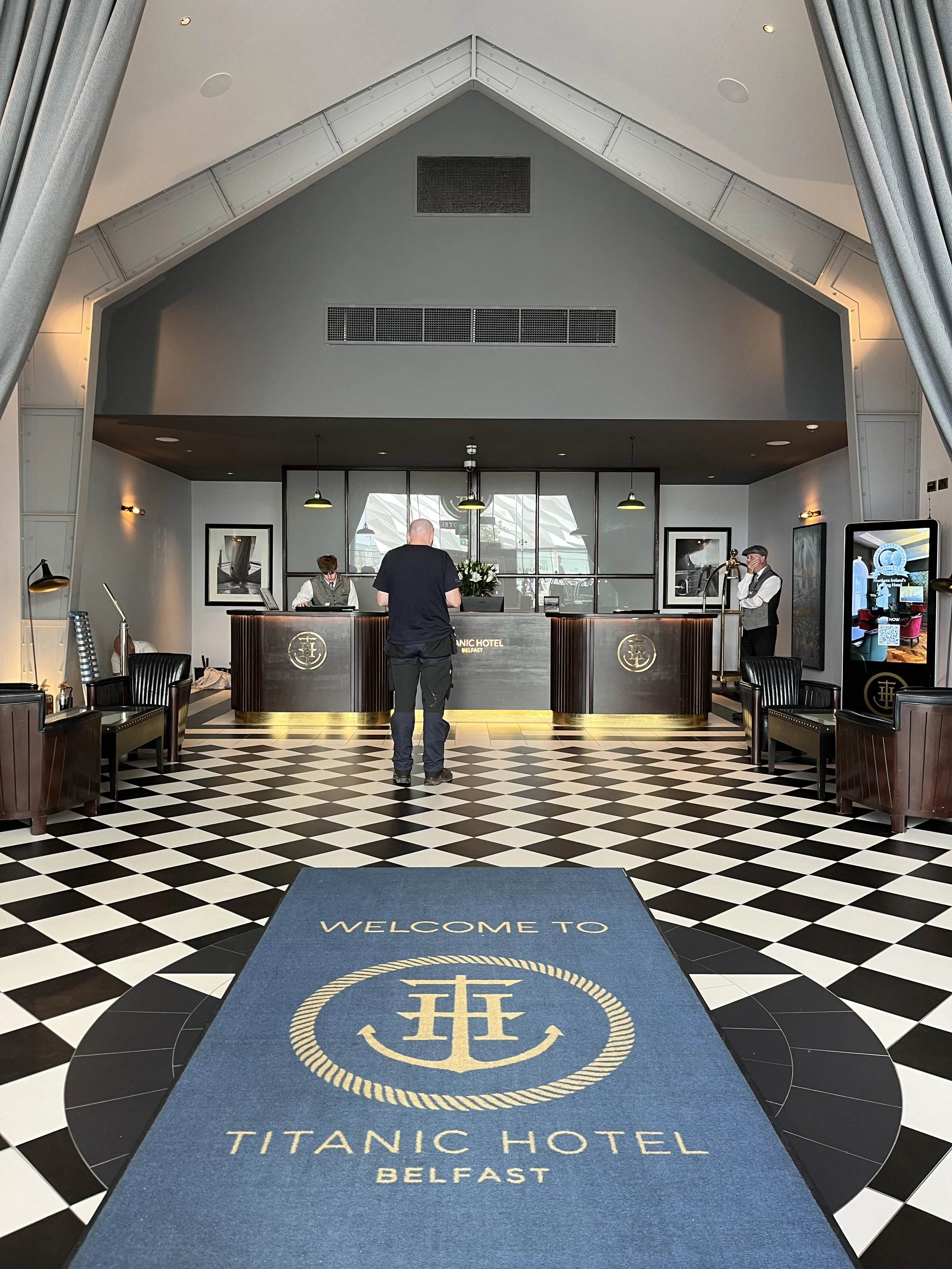 Vue de l'entrée et de la réception de l'hôtel Titanic à Belfast, comptoir en bois, carrelage à damiers, tapis bleu portant le logo de l'hôtel, ambiance luxe.
