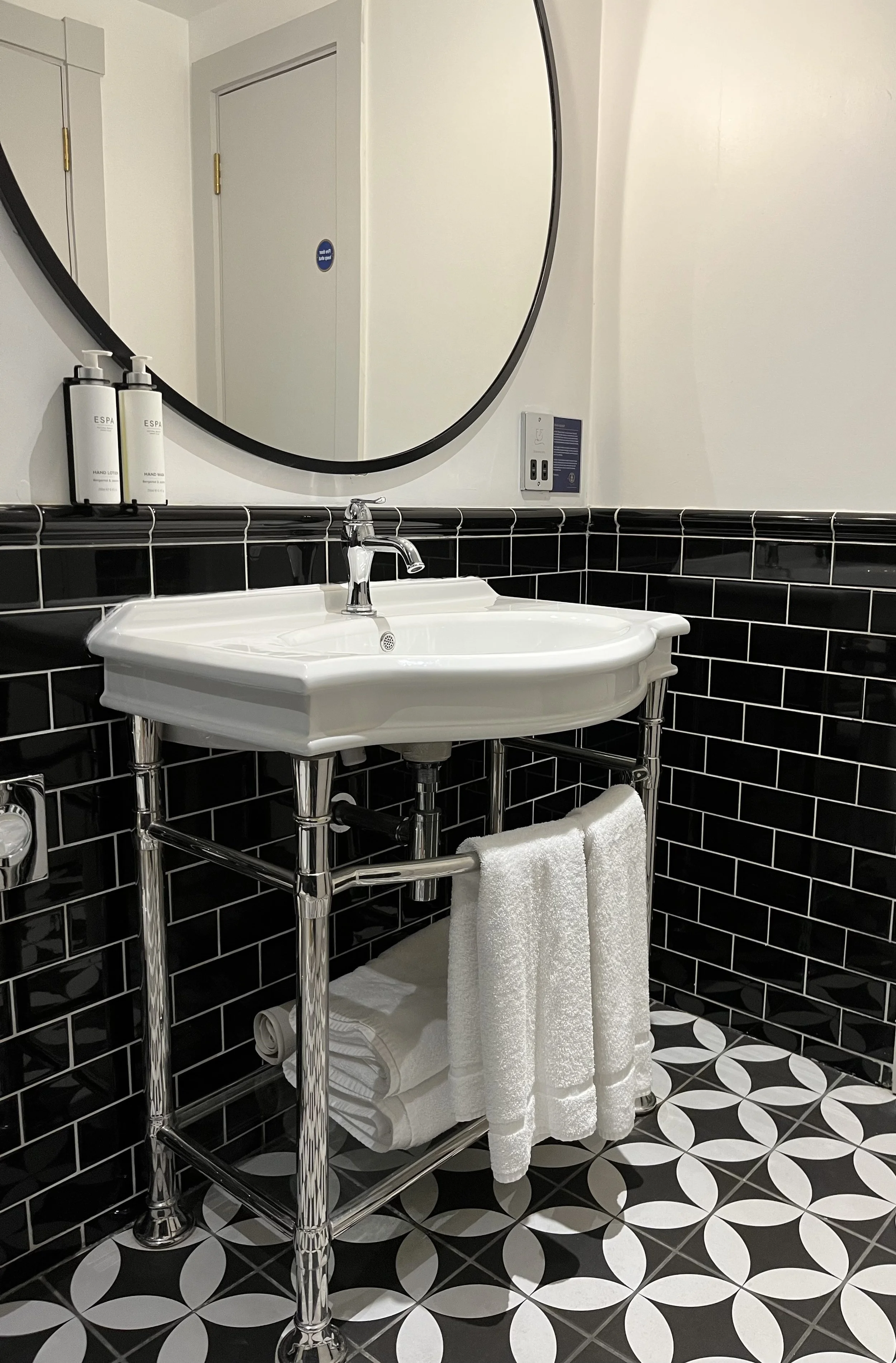 Magnifique salle de bains avec un lavabo blanc art déco, une grand miroir rond, un carrelage élégant noir et blanc, des serviettes de bains moelleuses, une cabine de douche au carrelage art déco noir avec une pluie de douche.