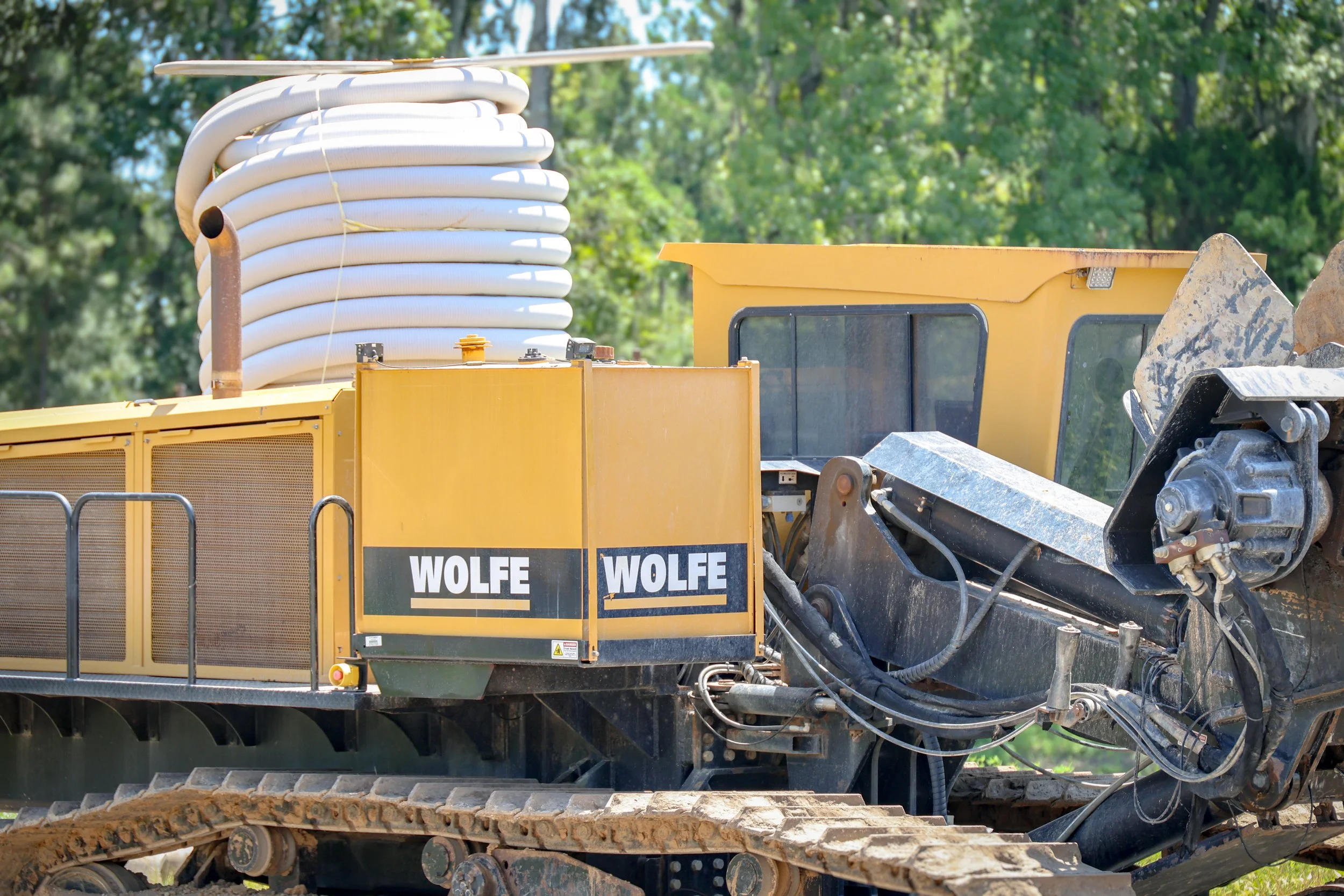 Close-up of a yellow construction vehicle with a coiled white hose on top and tracks at the bottom, set outdoors with green trees in the background. (Deepwater Dewatering machine -Hollywood, SC)