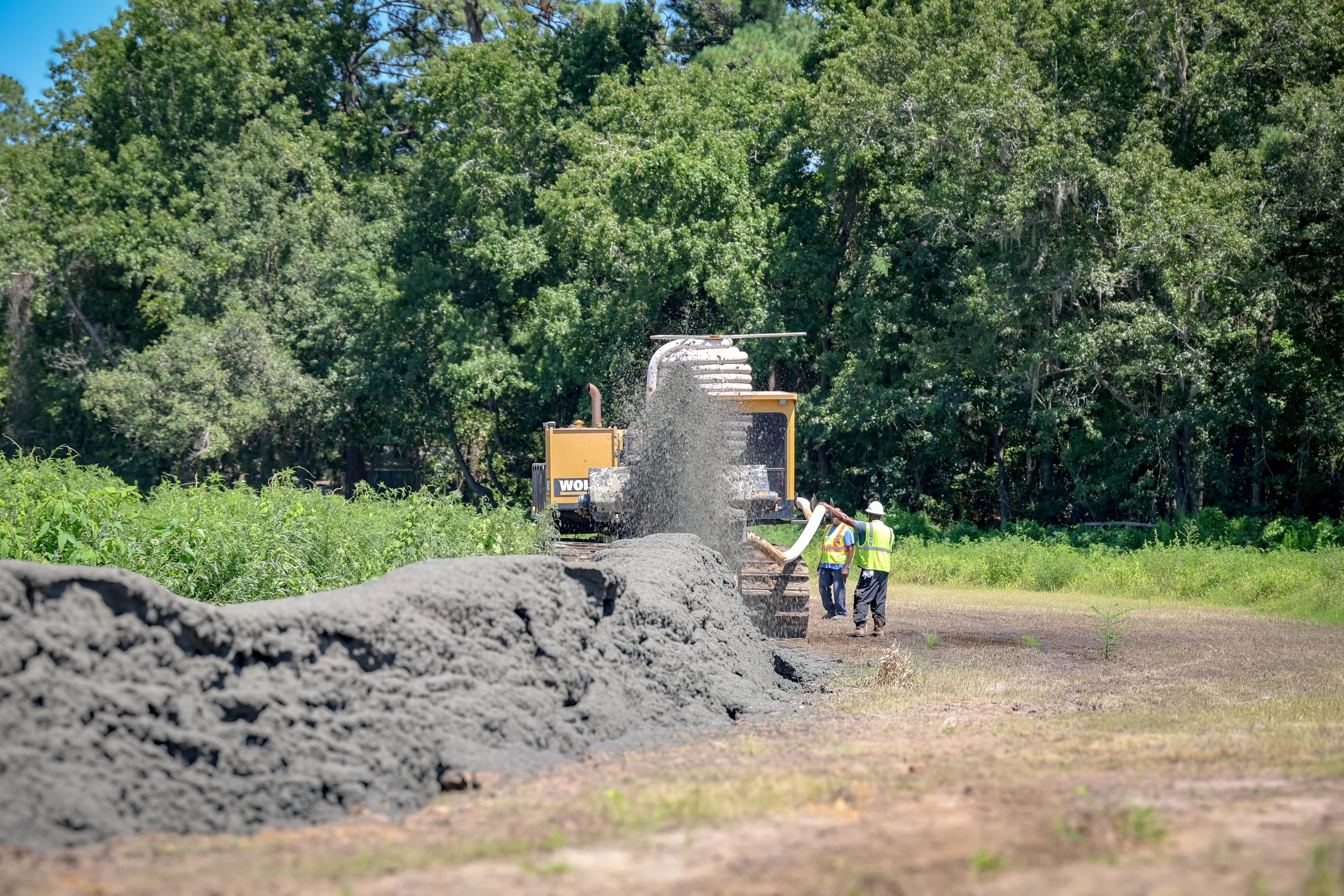 Construction workers in safety vests and helmets working on a road construction site surrounded by lush green trees, with a large pile of gravel in the foreground. (Deepwater Dewatering machine -Hollywood, SC)