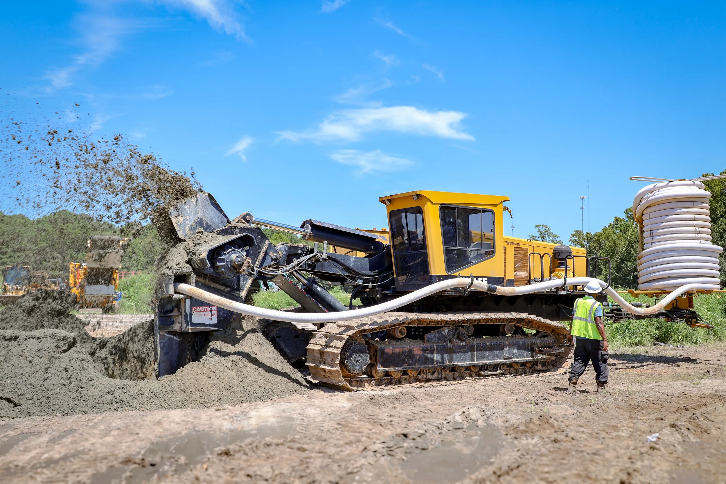 Construction site with a yellow bulldozer spreading dirt, a worker in a yellow vest observing, and a large coil of tubing on the right under a blue sky with some clouds. (Deepwater Dewatering machine -Hollywood, SC)