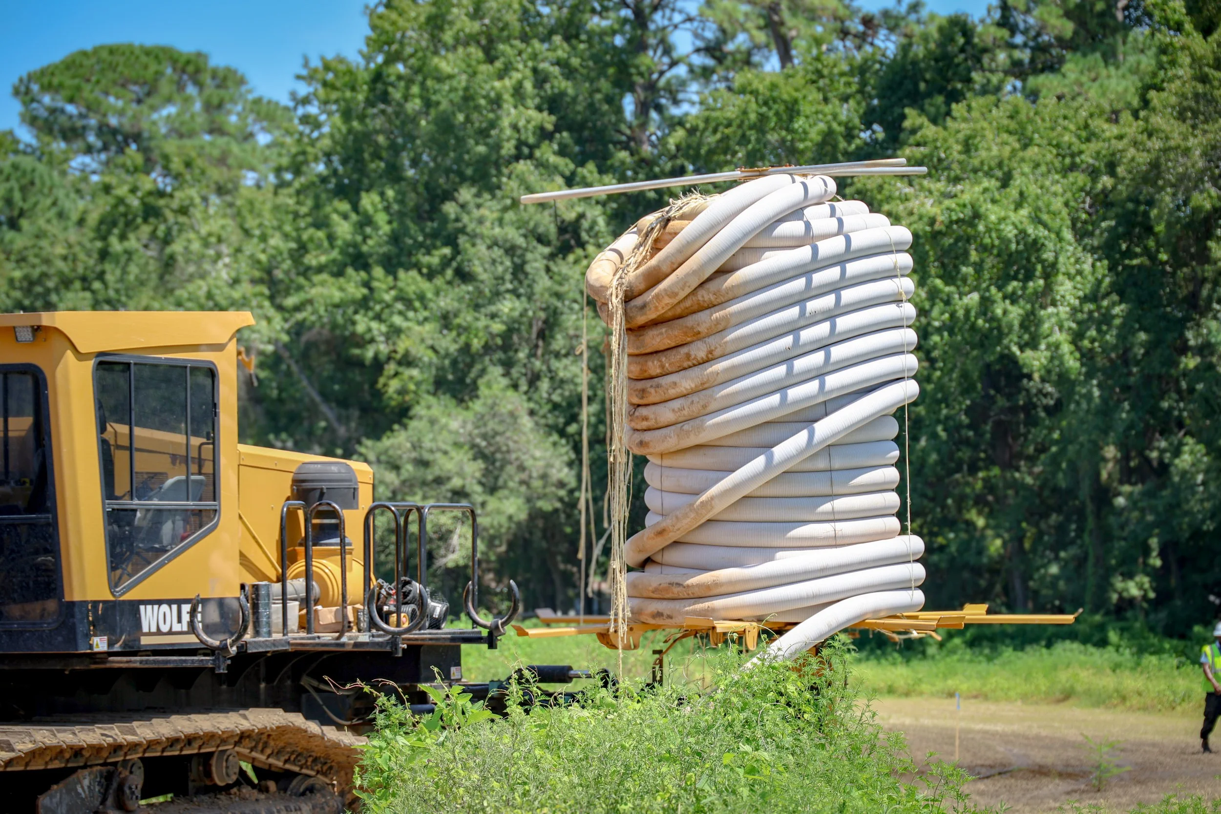 A large coil of white flexible piping is mounted on a yellow flatbed trailer attached to construction equipment with caterpillar tracks. The background features green, leafy trees and a clear blue sky. (Deepwater Dewatering machine -Hollywood, SC)