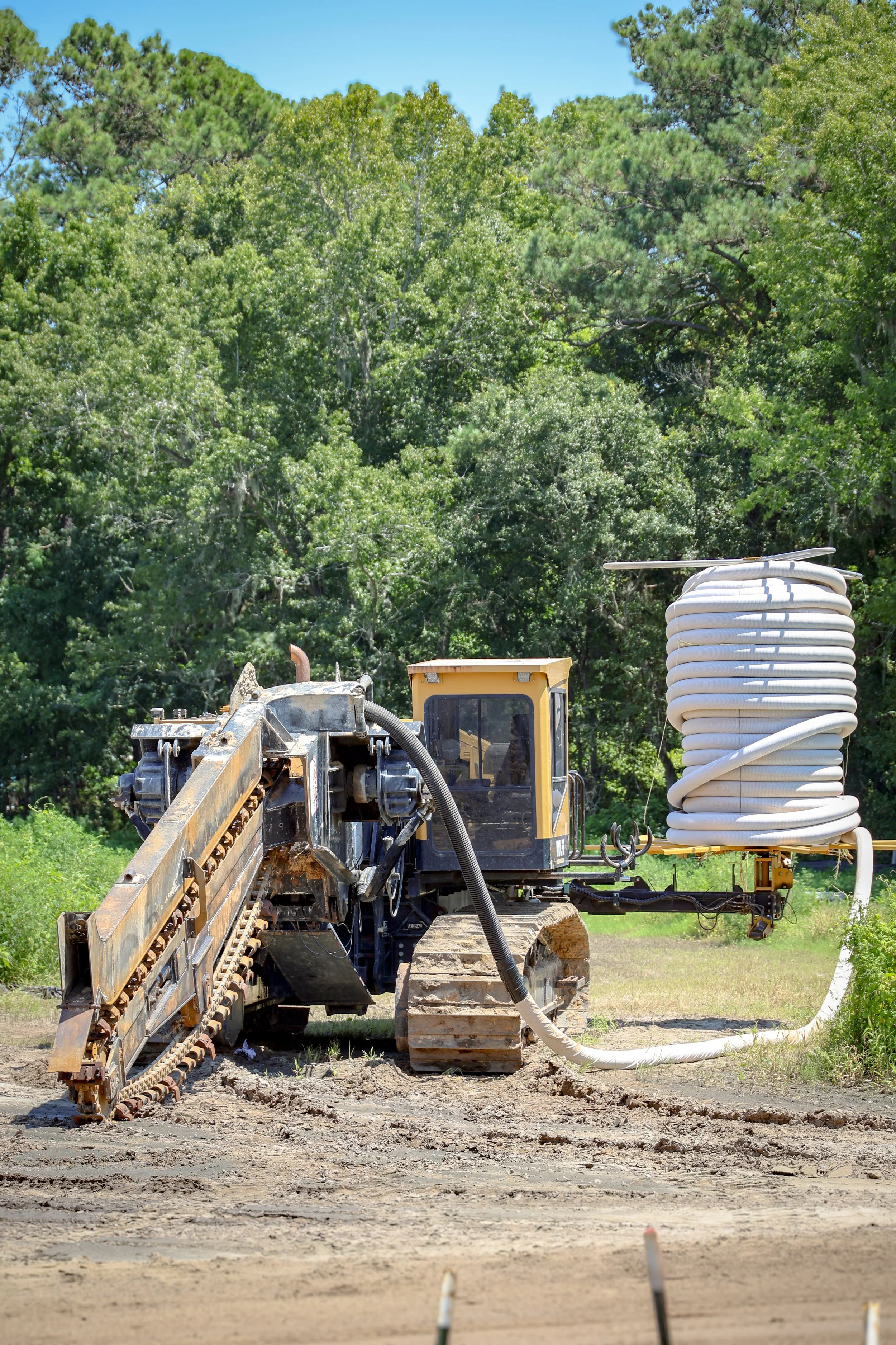 A construction site featuring a bulldozer with caterpillar tracks and a large coil of white tubing mounted on a yellow frame, set against a backdrop of lush green trees and a bright blue sky. (Deepwater Dewatering machine -Hollywood, SC)