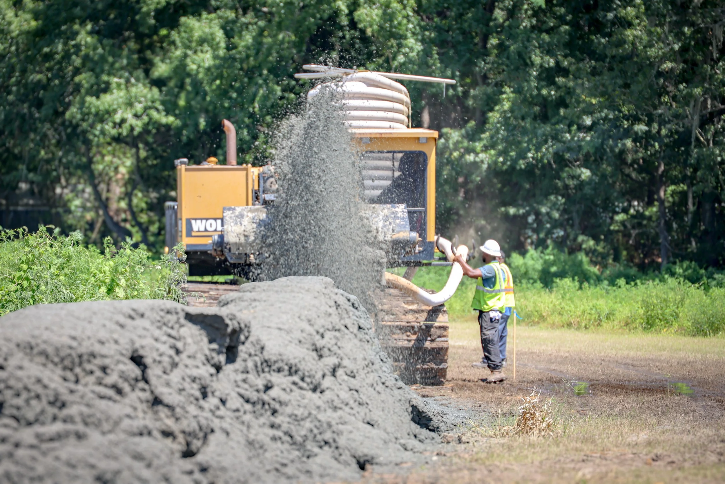 Construction workers using a machine to spread gravel on a road with greenery in the background. (Deepwater Dewatering machine -Hollywood, SC)