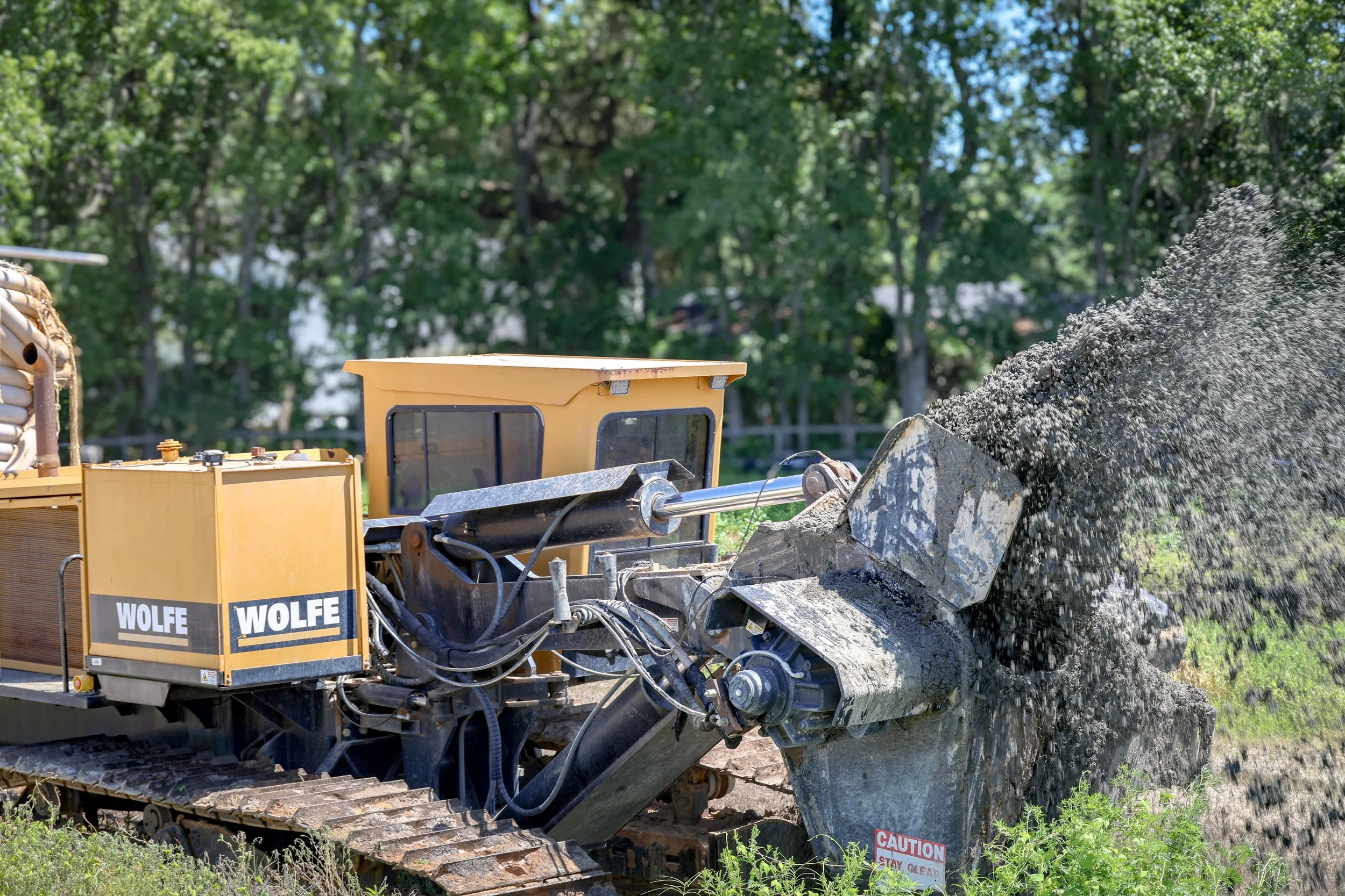 A large yellow industrial construction machine breaking large concrete rocks with water spraying out. There is a wooded area in the background. (Deepwater Dewatering machine -Hollywood, SC)