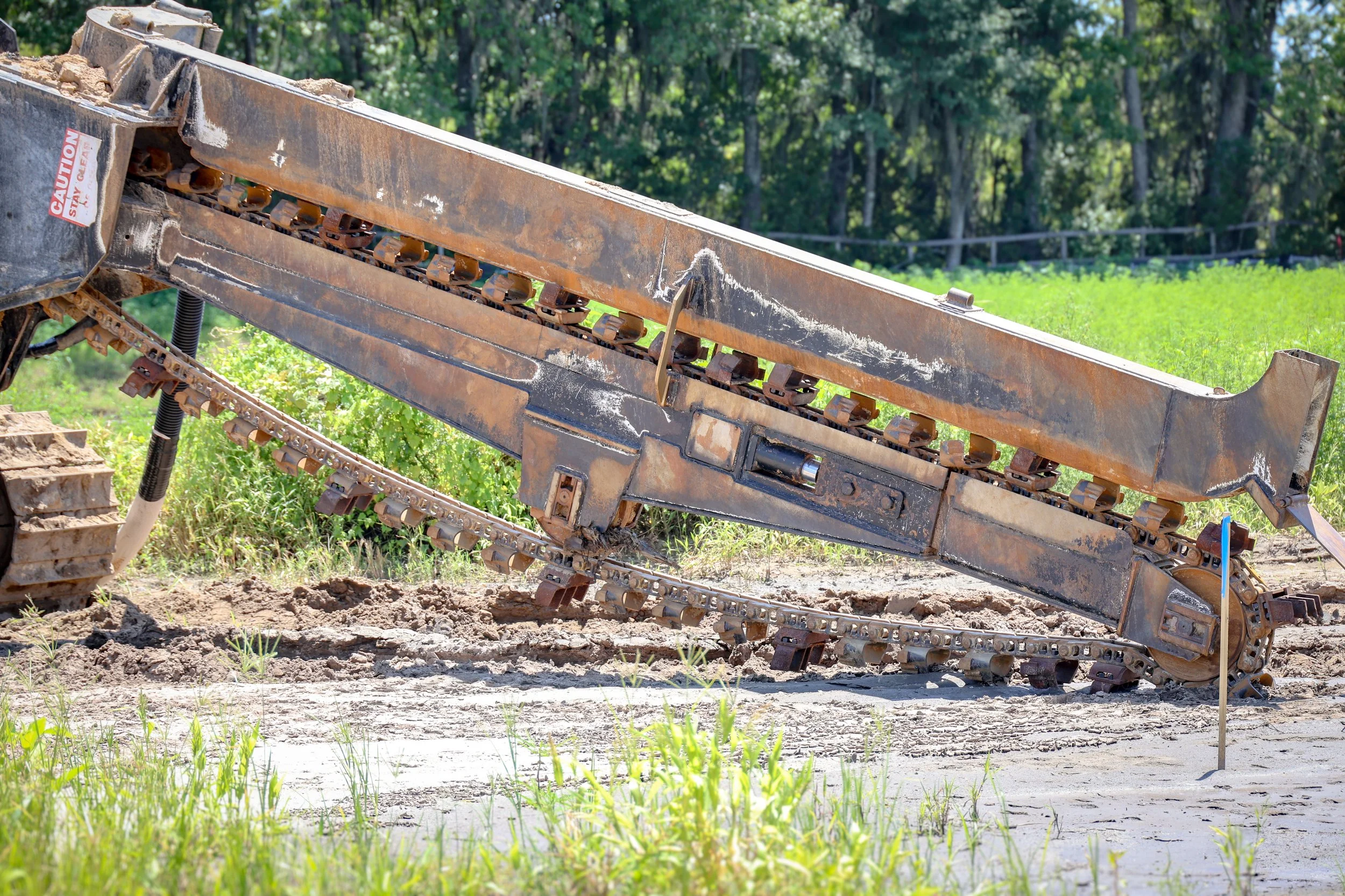 A large, rusty piece of construction equipment in a dirt and grassy outdoor area with green trees and a wooden fence in the background.