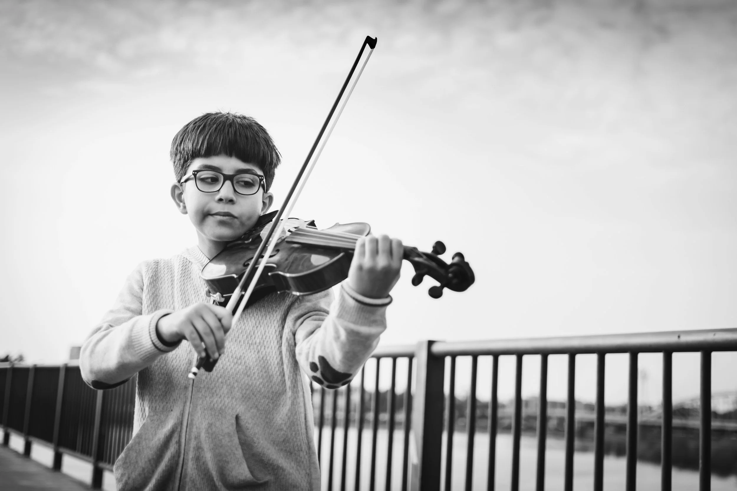 A young boy wearing glasses playing the violin outdoors, with buildings and a fence in the background.
