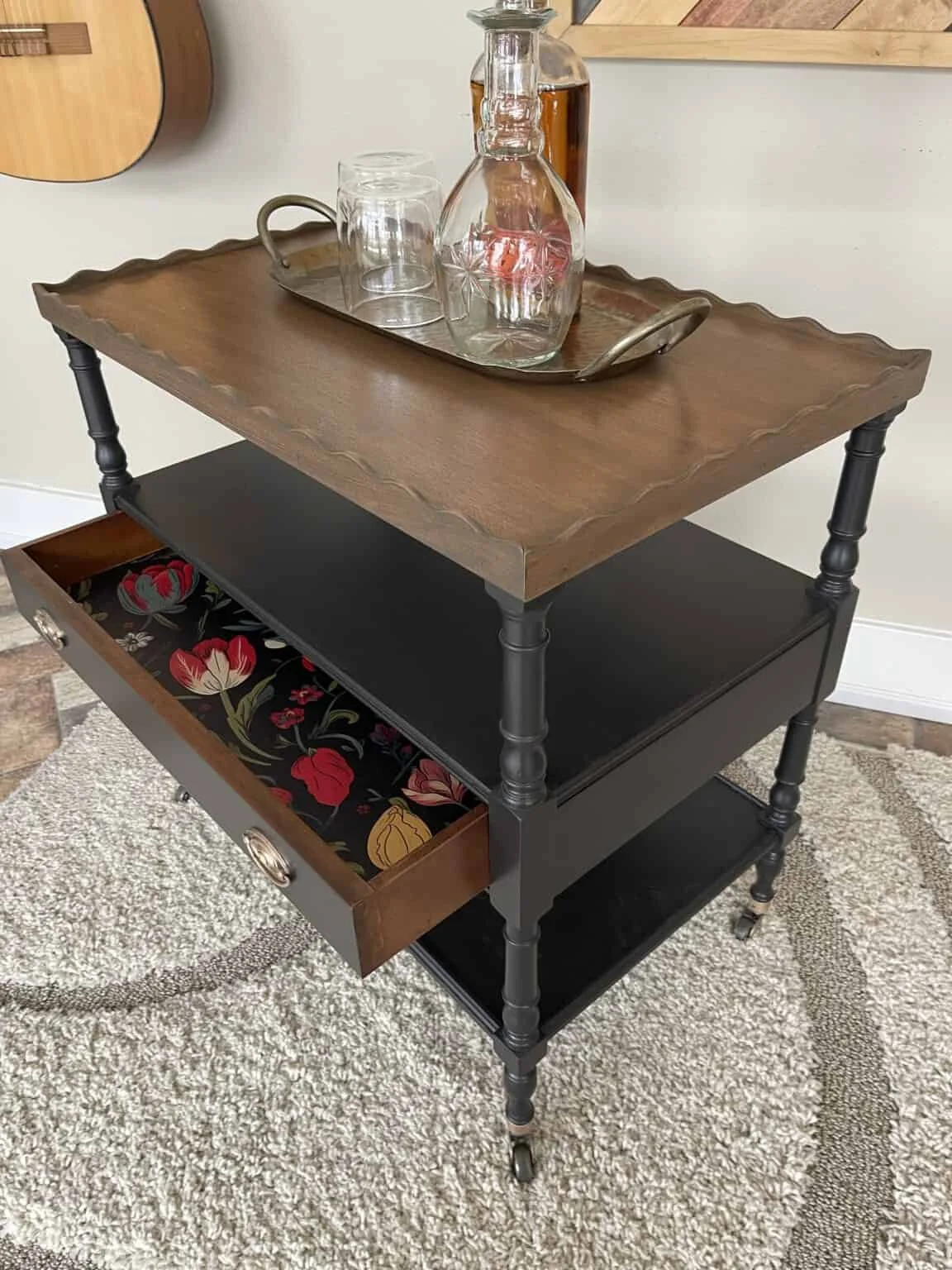 A three-tier rolling bar cart with a wooden top, black middle shelf, and a drawer with floral interior, holding glassware and bottles.