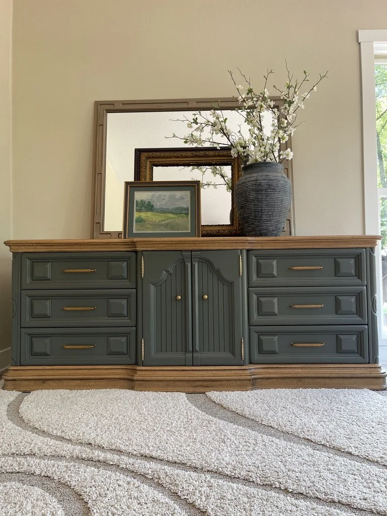 A dark green wooden sideboard with gold handles and a light wood top, featuring a large dark vase with white branches and flowers, two framed mirrors, and a small landscape painting on top, with a white textured rug and a window with sunlight in the background.