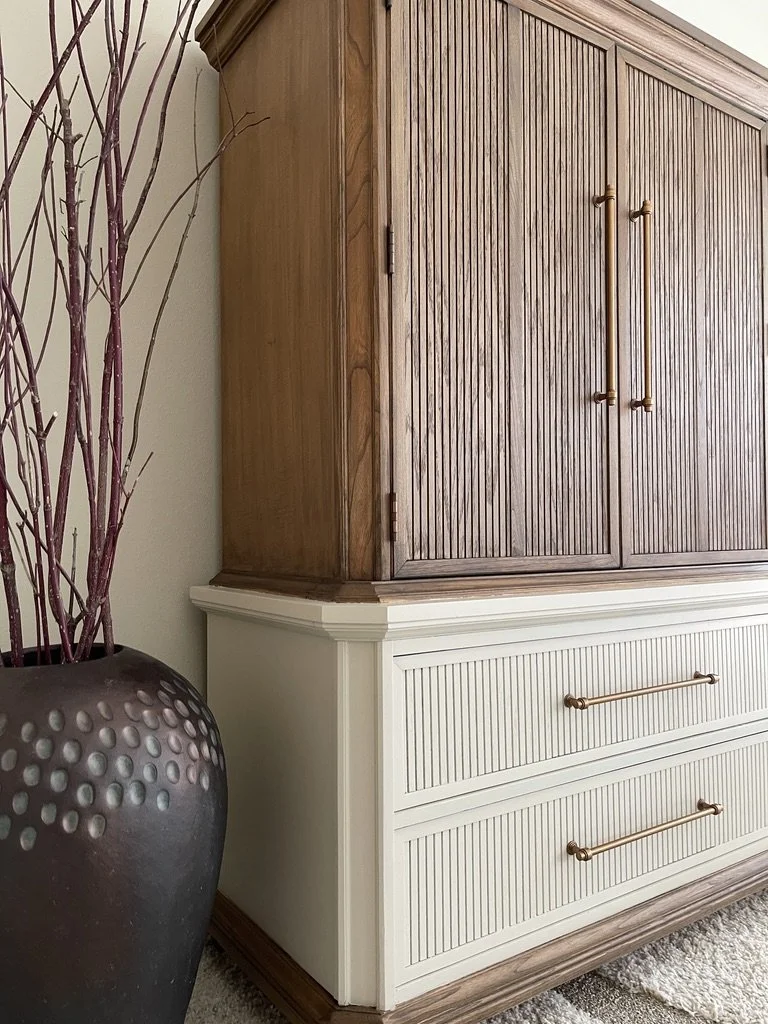 Close-up of a corner cabinet with wood-paneled upper section and white lower drawers with metallic handles, next to a tall, dark vase with decorative dots containing branches, on a carpeted floor.