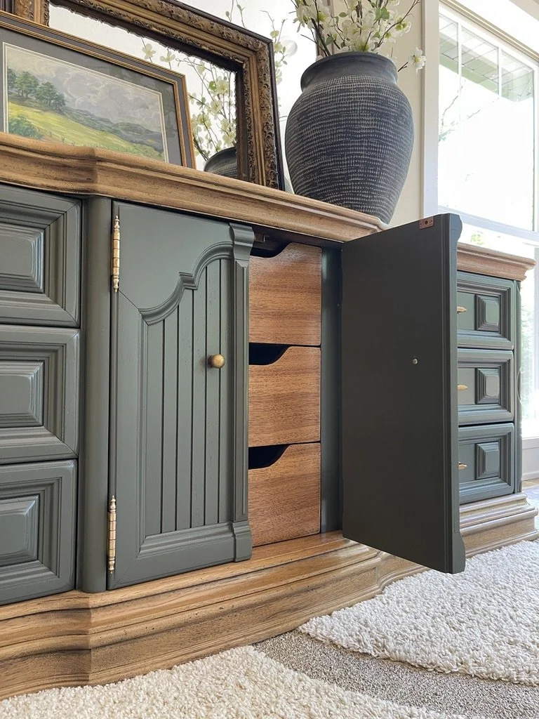 A close-up of a wooden and painted cabinet with open doors revealing drawers inside, topped with a large Potter-style black vase with white flowers, a framed picture, and a window in the background.