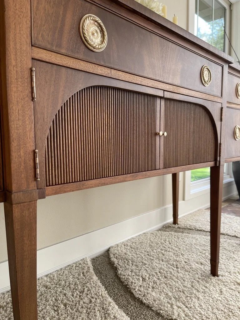 Close-up of a vintage wooden sideboard with decorative metal medallions and ribbed sliding doors, placed in a room with a window and carpeted floor.