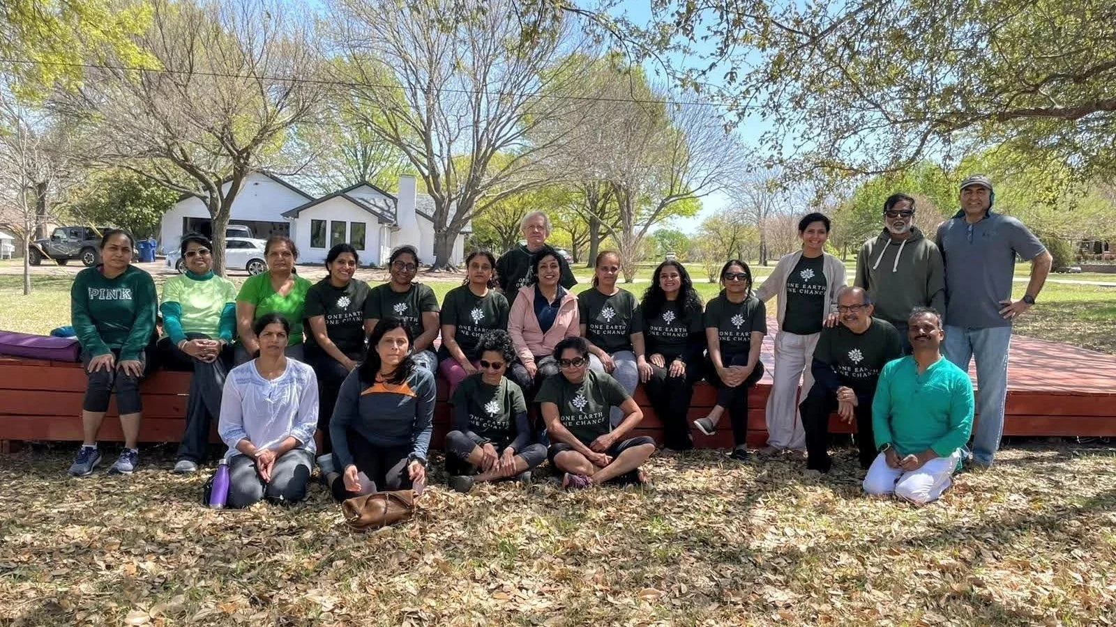 Group of people outdoors sitting and standing on a wooden platform in a park with trees and houses in the background.
