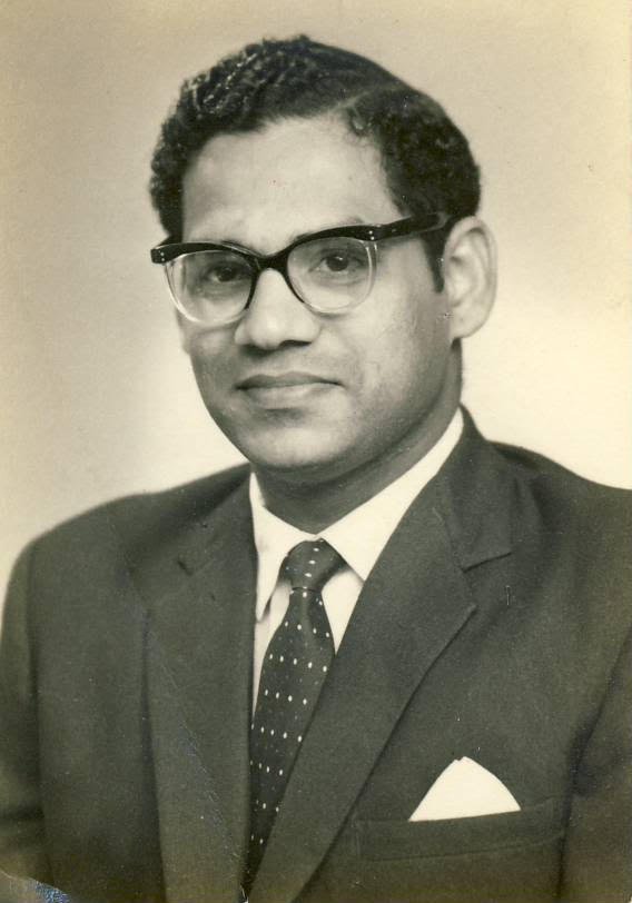 Black and white portrait of a man wearing a suit, tie, and glasses, facing the camera with a serious expression.